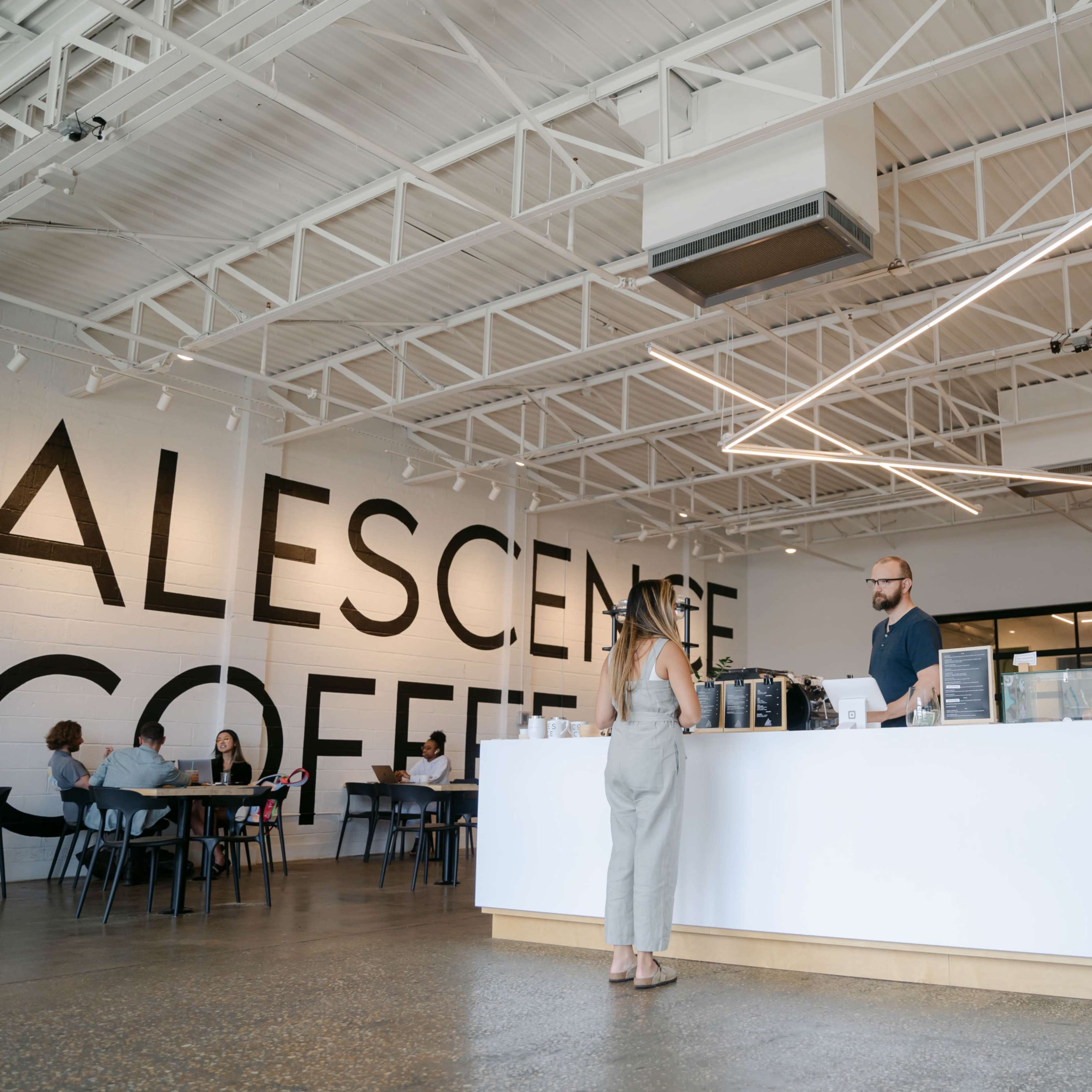 A coffee shop features a large white counter with a barista serving a customer, while several patrons sit at tables in the background.