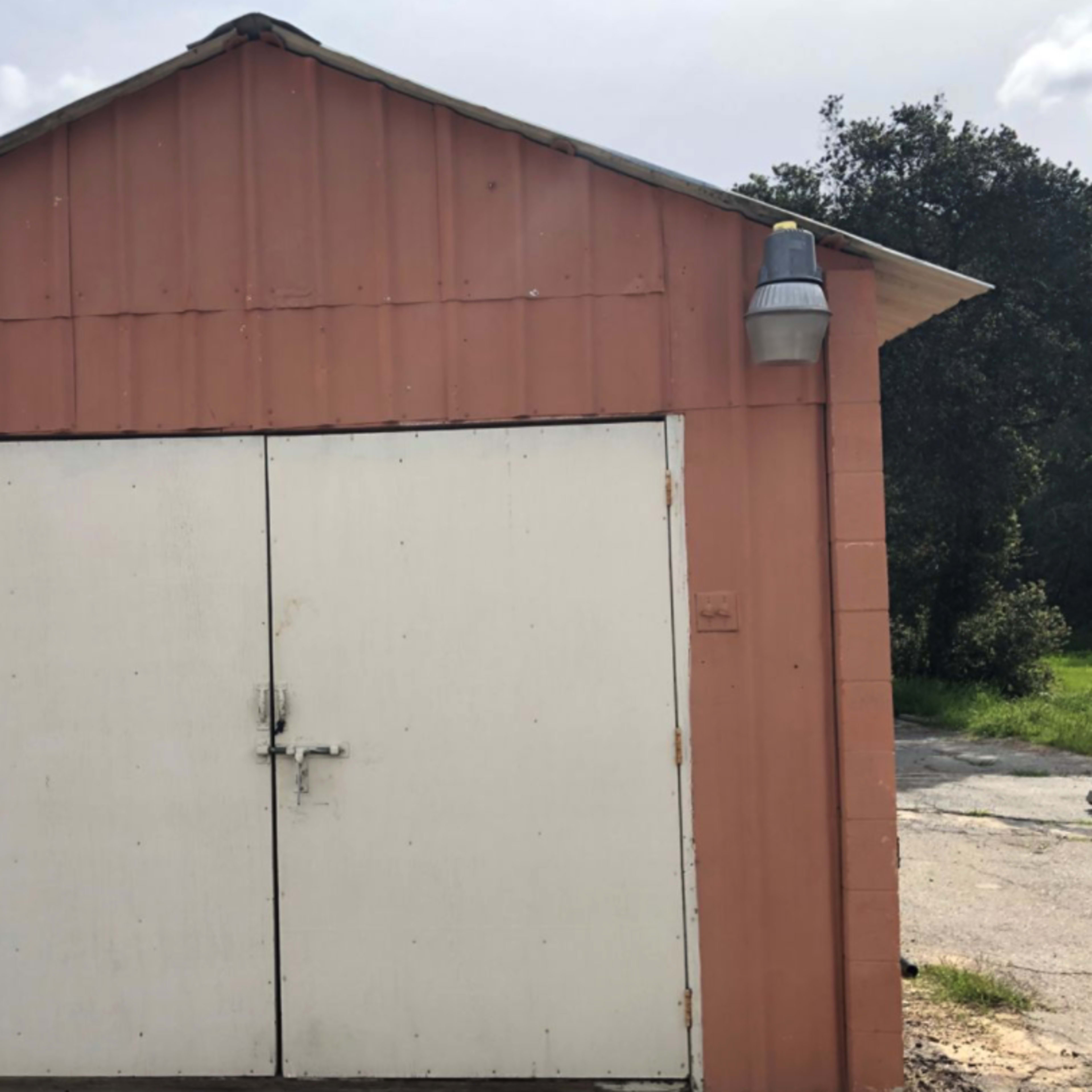 A small, orange shed with a white double door, set against a backdrop of greenery under a cloudy sky.
