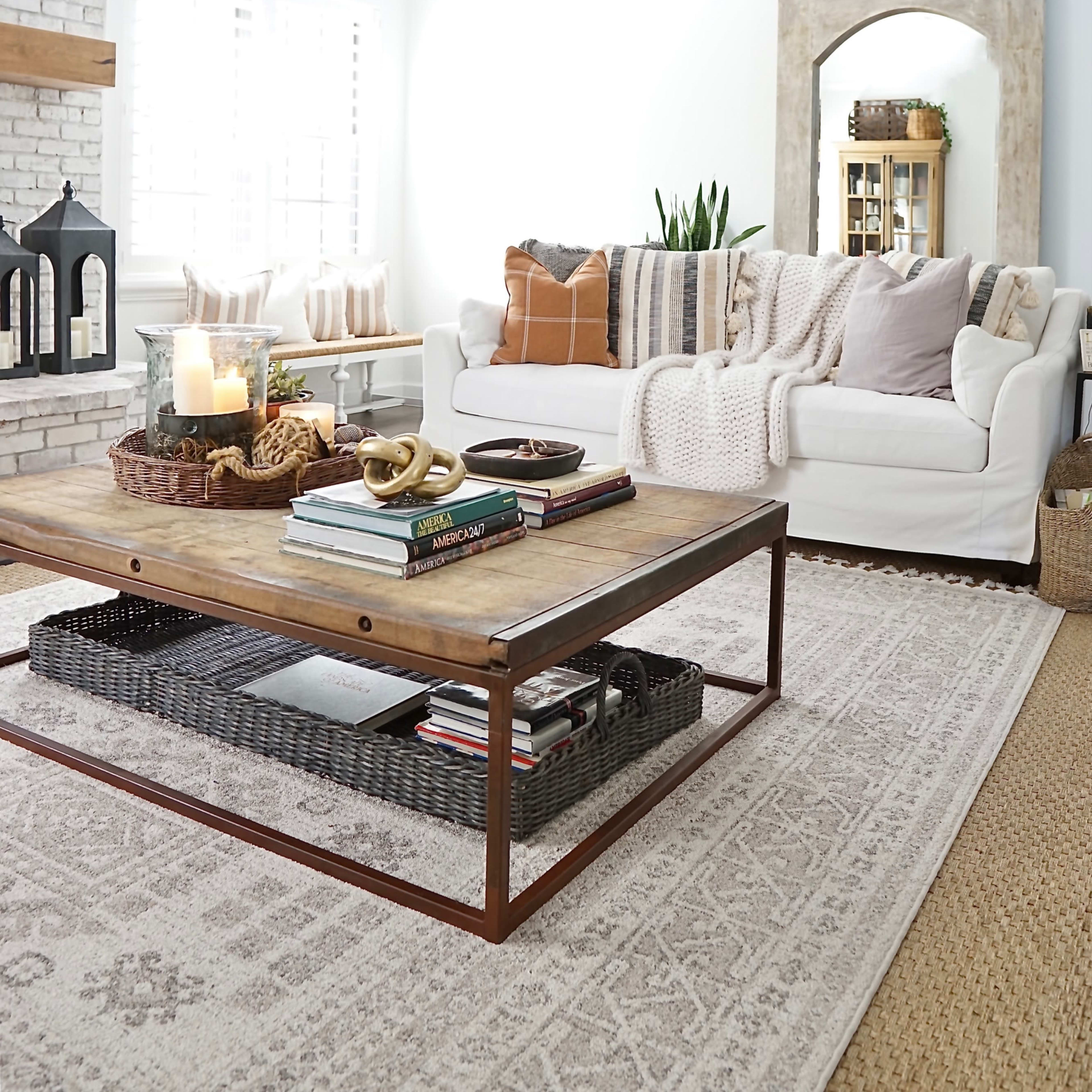 A bright living room features a white sofa adorned with throw pillows, a wooden coffee table with stacks of books, and a patterned area rug.