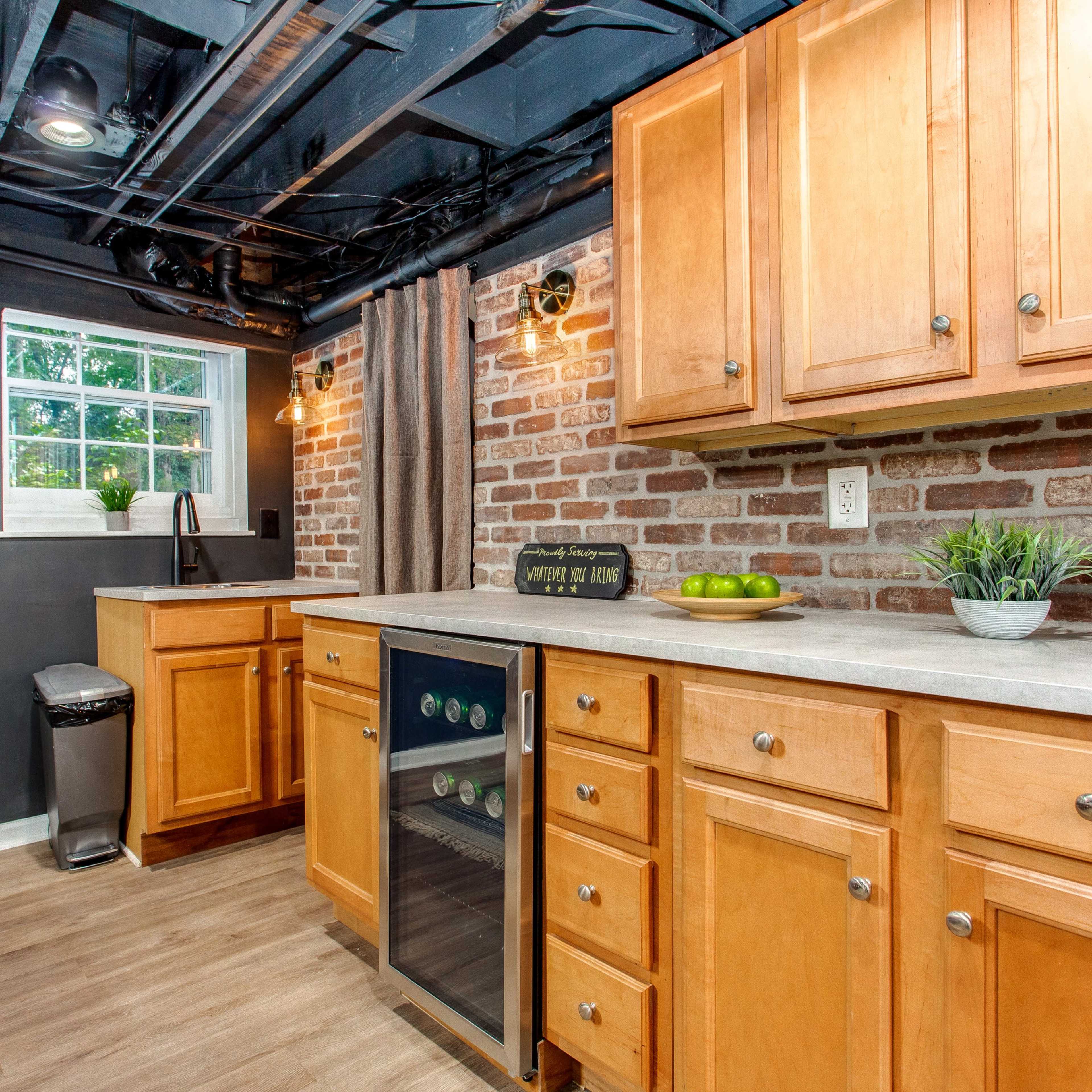 A modern kitchen with wooden cabinets, a stone countertop, and a small wine refrigerator, against a backdrop of exposed brick walls.