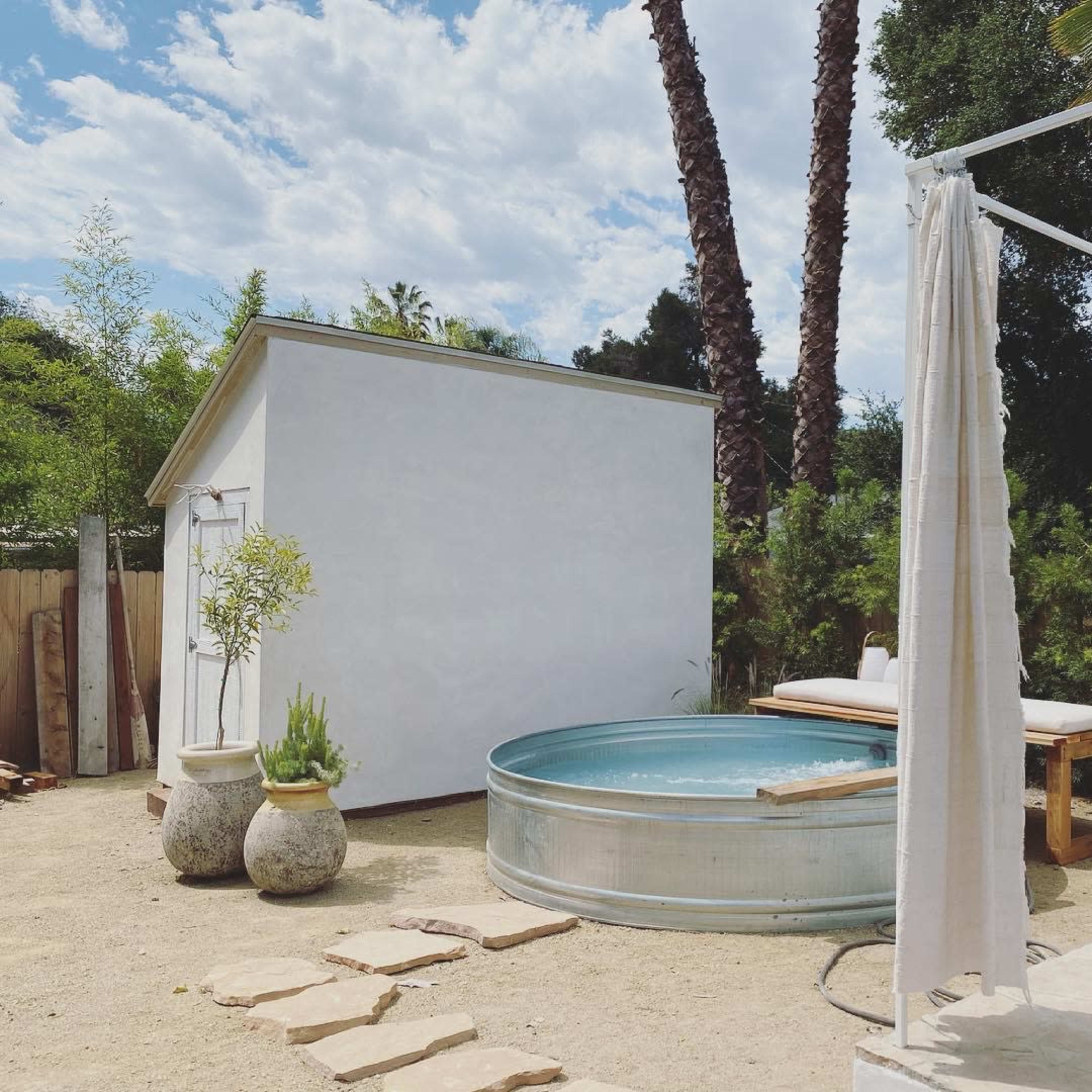 A round metal tub is situated on a sandy patio surrounded by palms and a white shed, with light-colored curtains draping nearby.