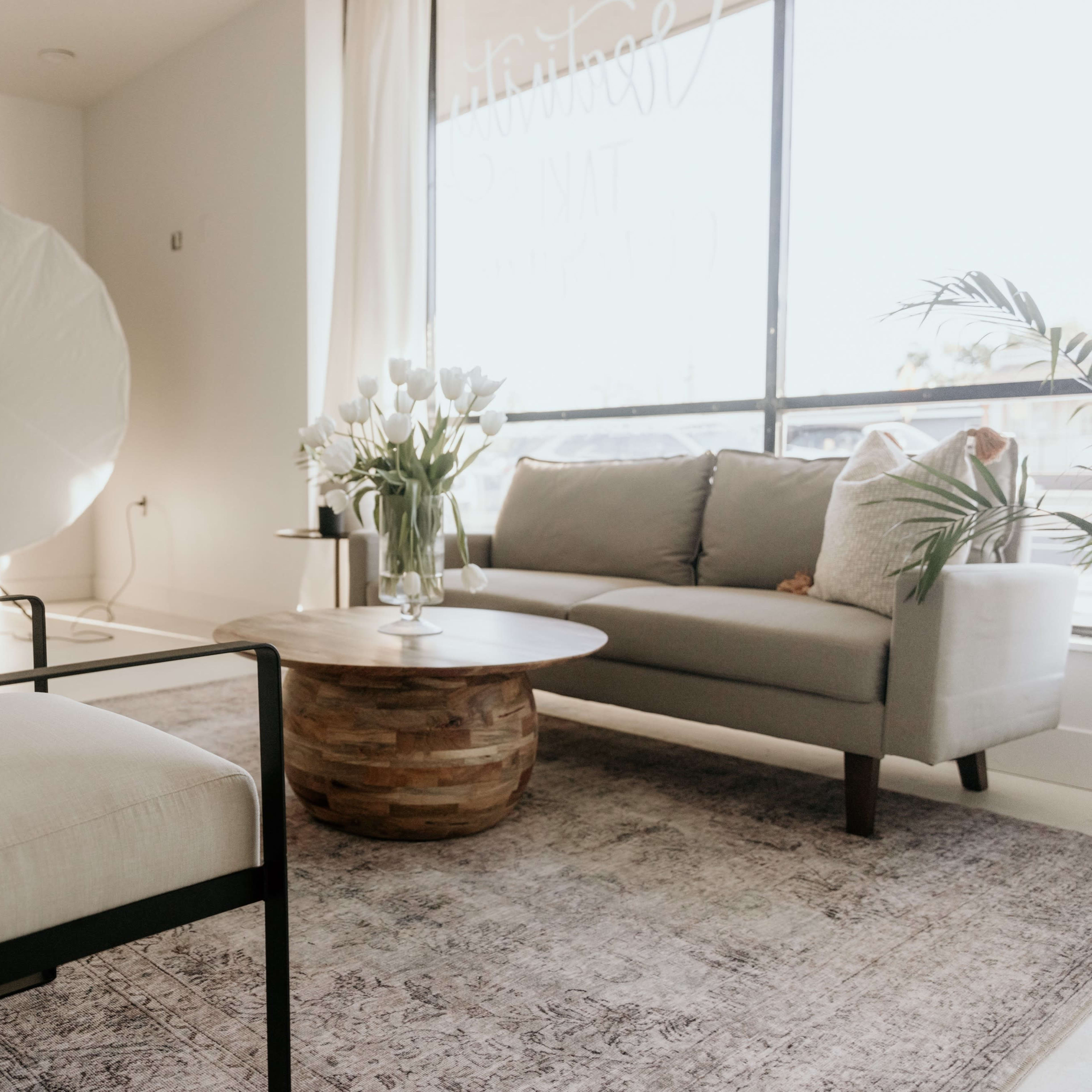 A modern, minimalistic living room with a gray sofa, two light-colored chairs, a round wooden coffee table, and a large light reflector in the background.