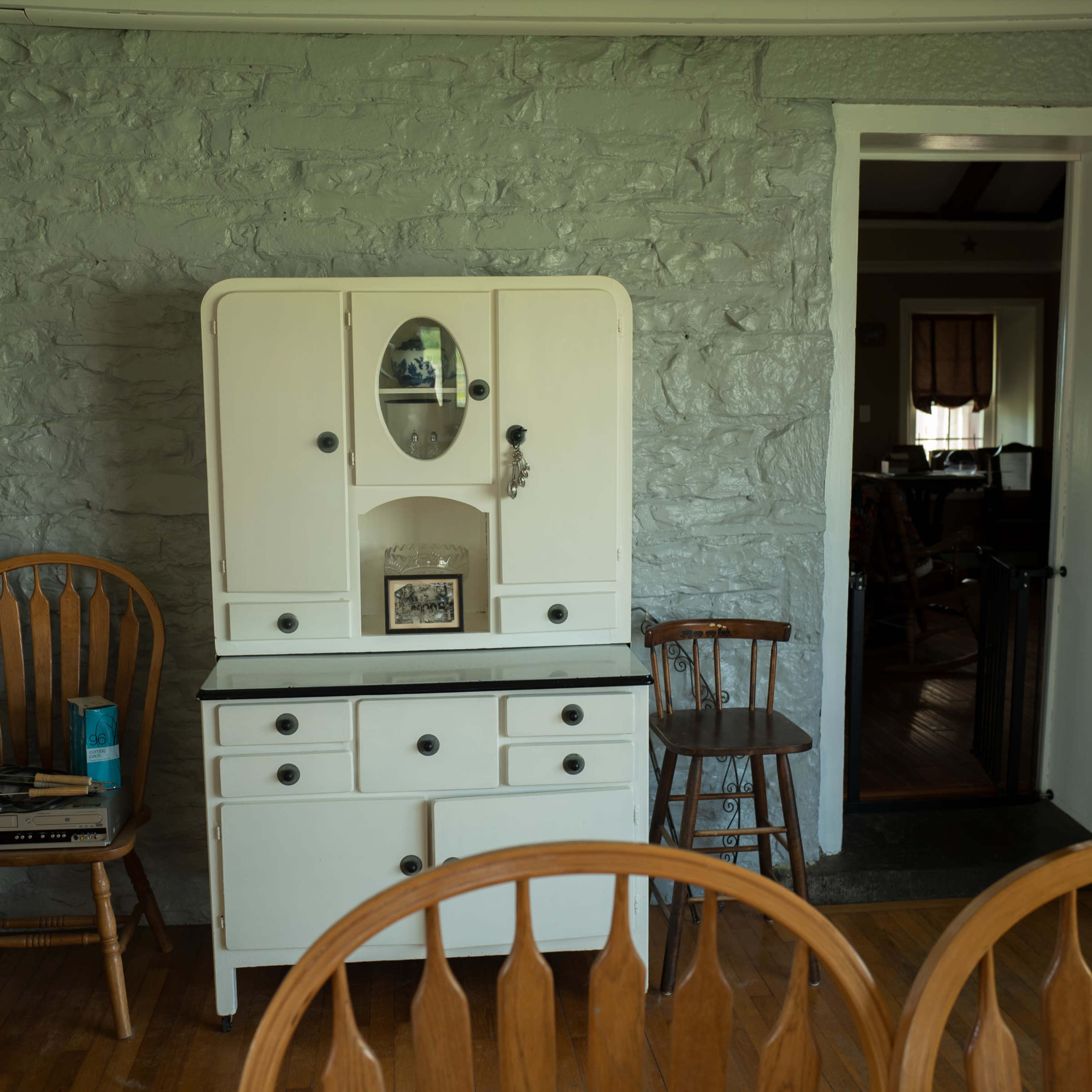 A dining area with a stone gray wall, a white cabinet, wooden chairs, and an open doorway leading to another room.