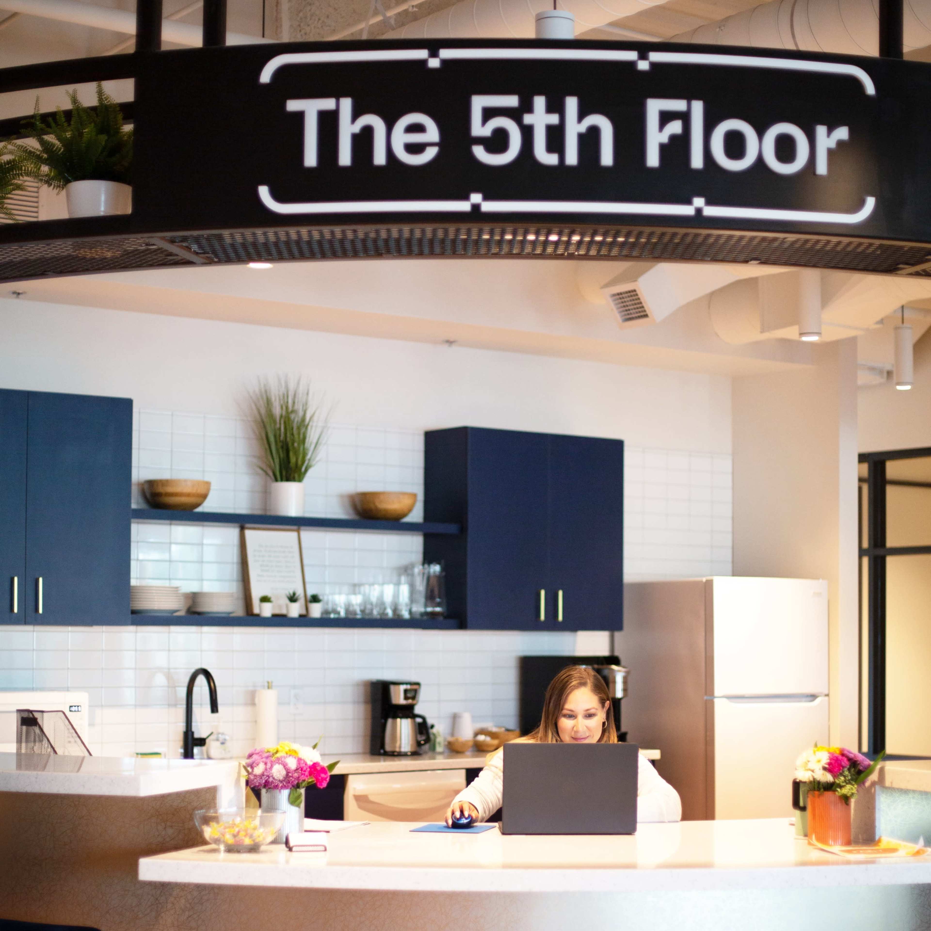 A woman works on a laptop at a modern reception desk in an office space labeled "The 5th Floor."