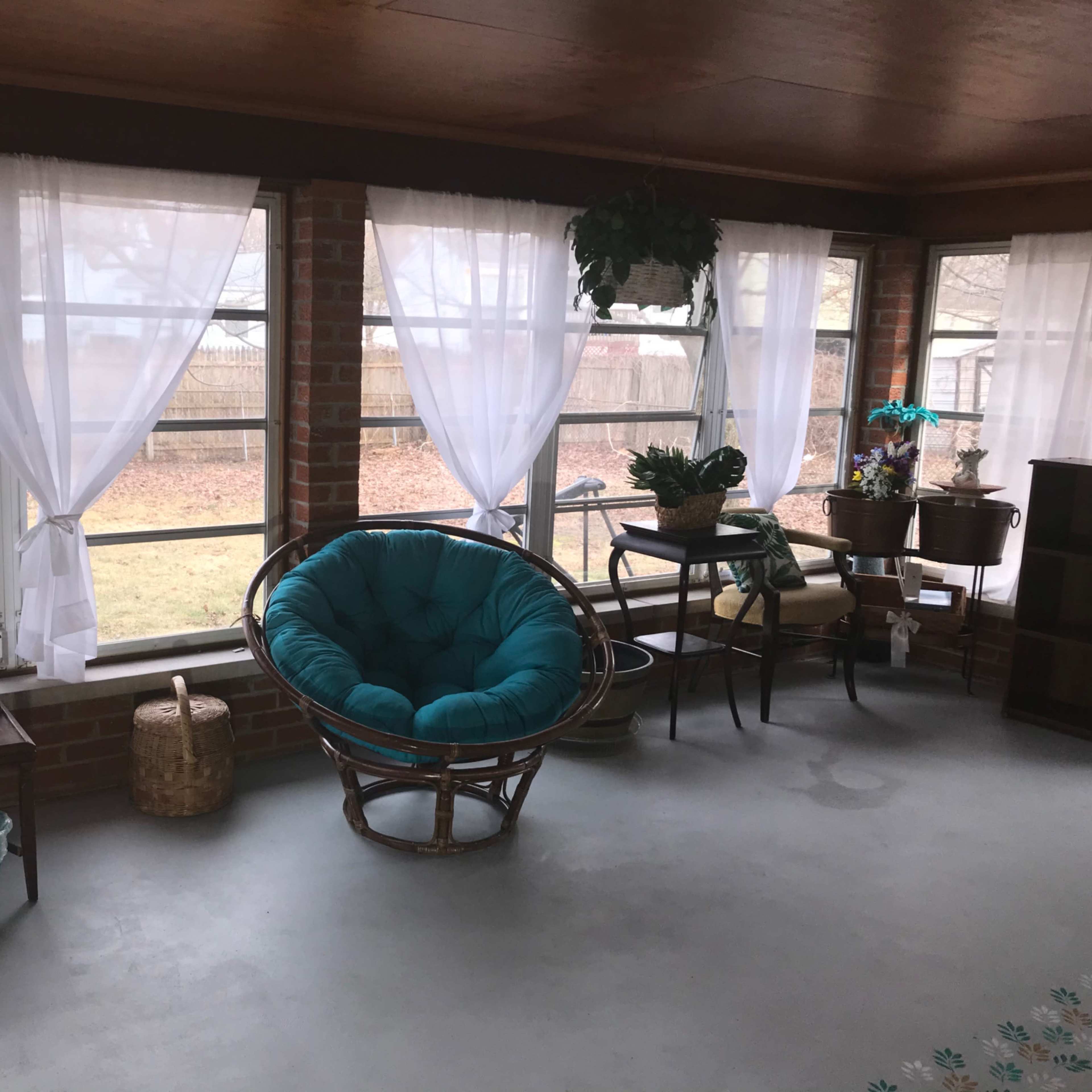 A sunroom with large windows, featuring a round turquoise chair, a couple of tables, and decorative plants.