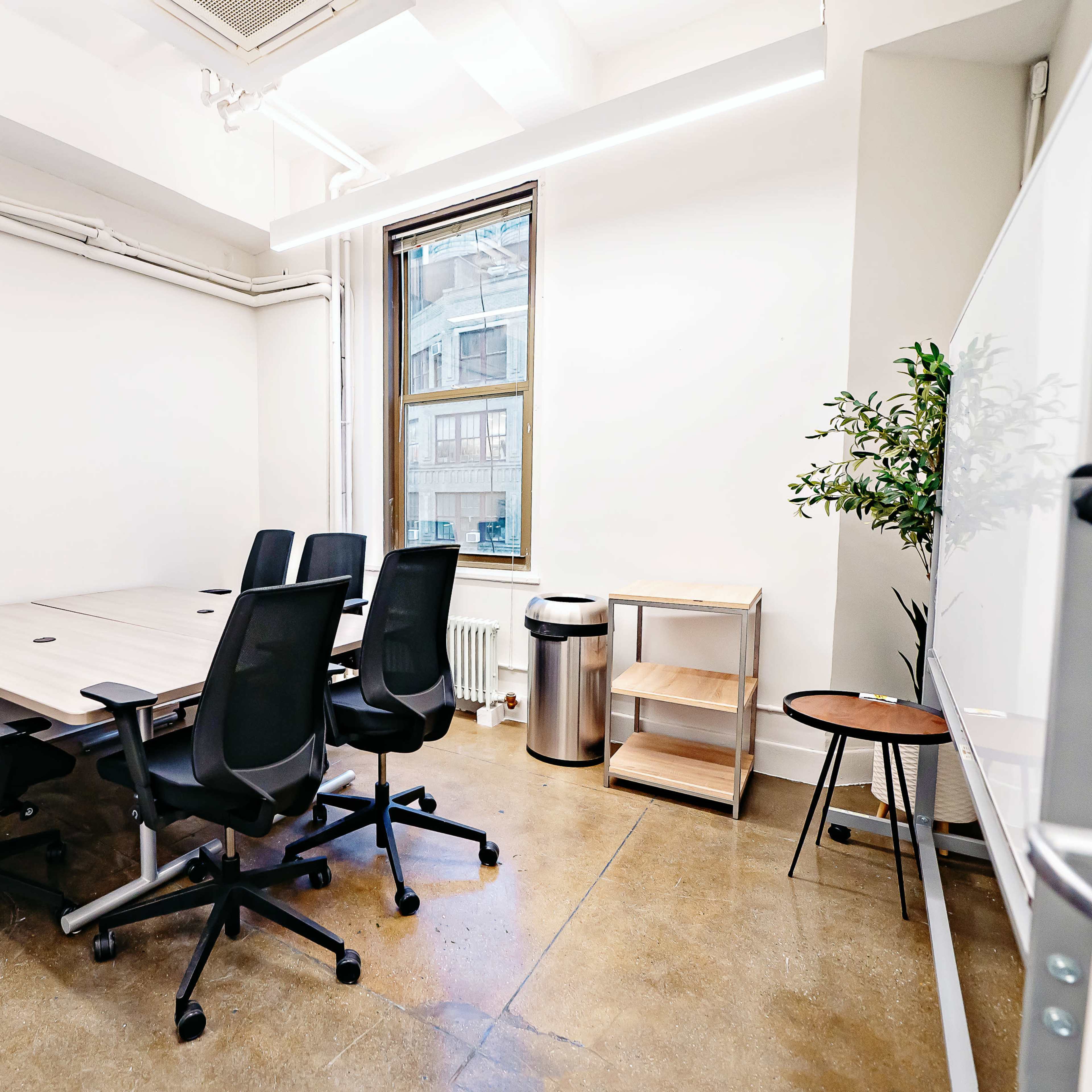 A modern conference room features a large table surrounded by six black office chairs, with a window, a small plant, and a waste bin in the corner.