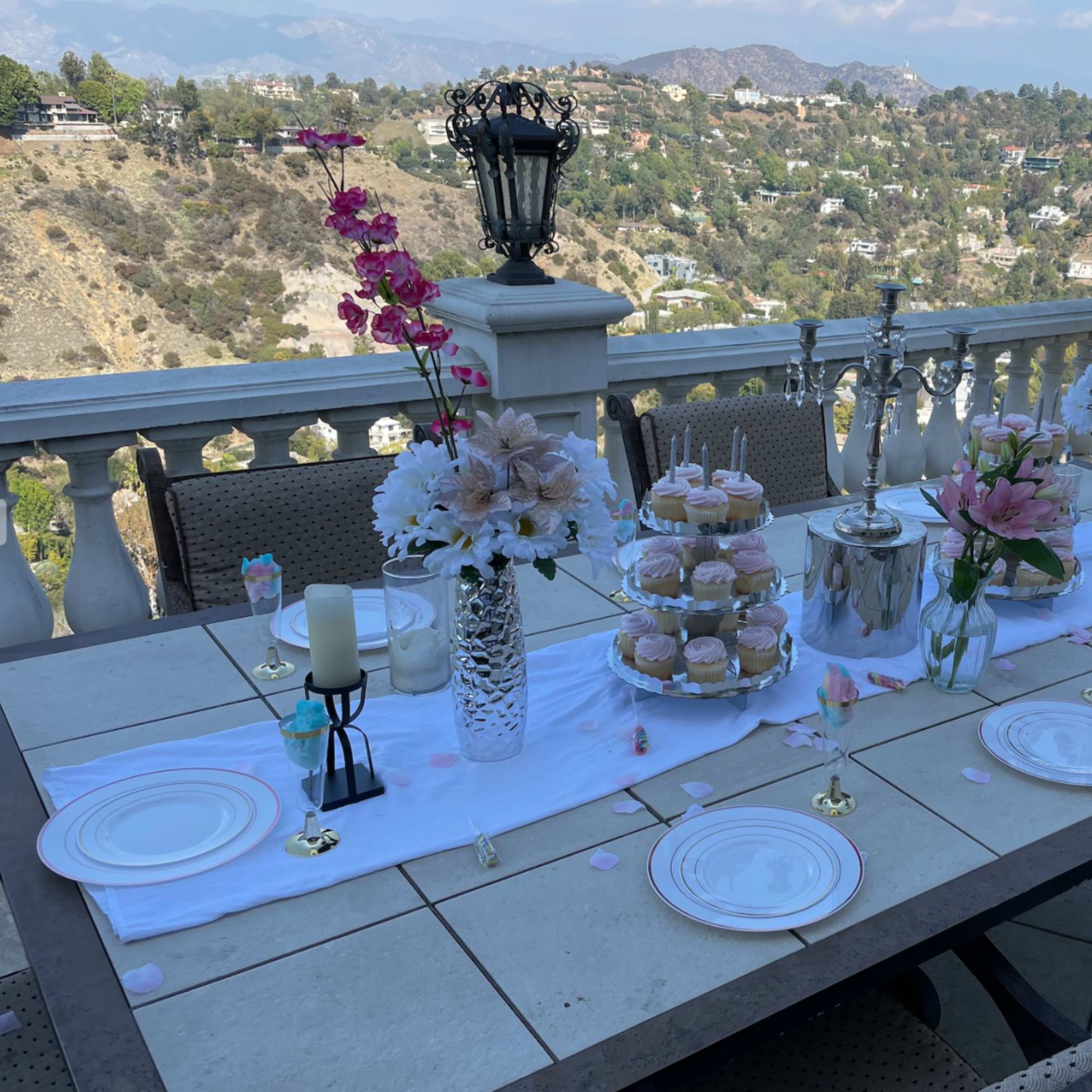 A decorated outdoor table is set with elegant tableware, floral arrangements, and cupcakes against a backdrop of hills and distant mountains.