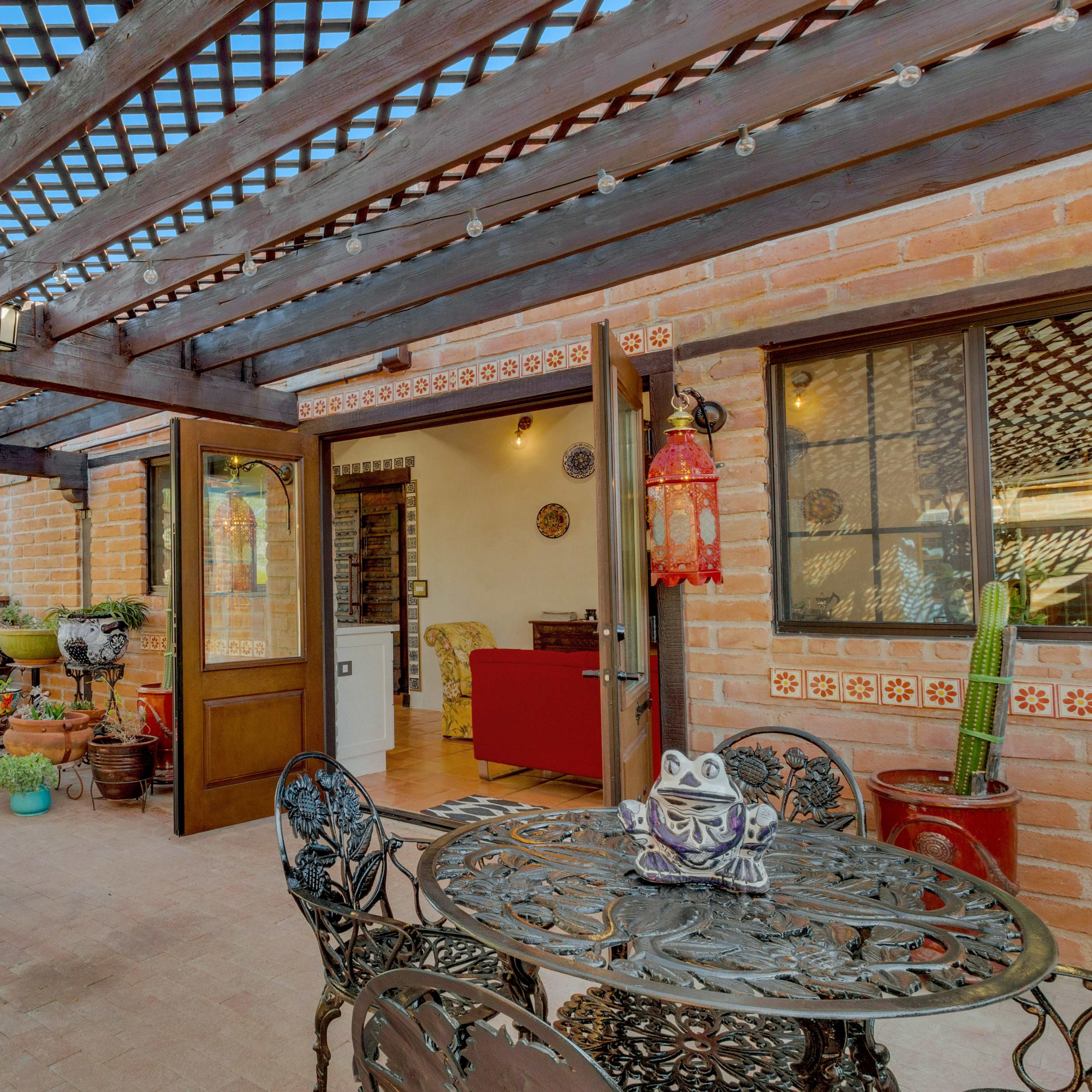 A terrace area with a wrought iron table and chairs, surrounded by potted plants, and features a wooden pergola overhead.