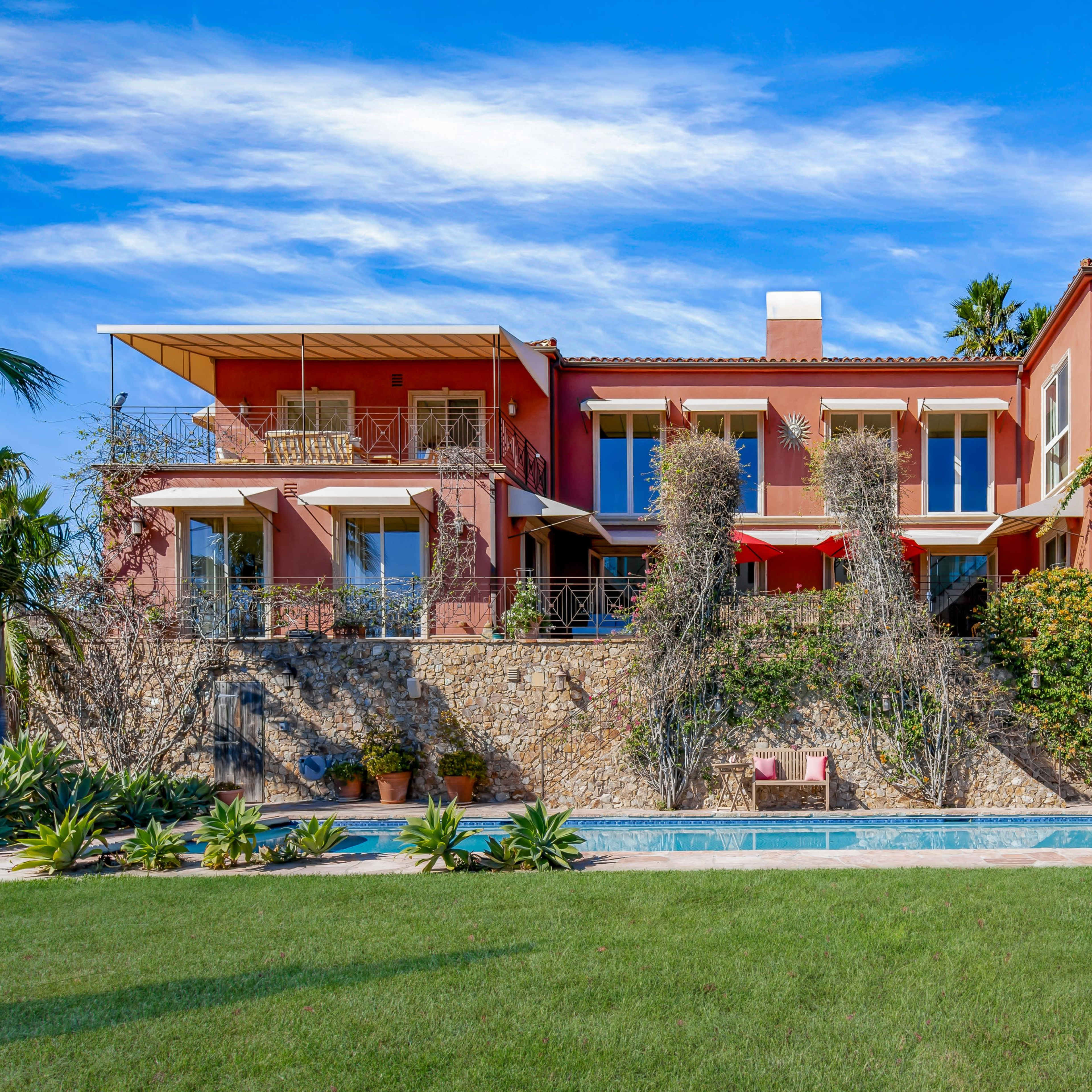 A large, multi-level red villa with balconies sits alongside a swimming pool and a green lawn, surrounded by palm trees and vibrant landscaping.