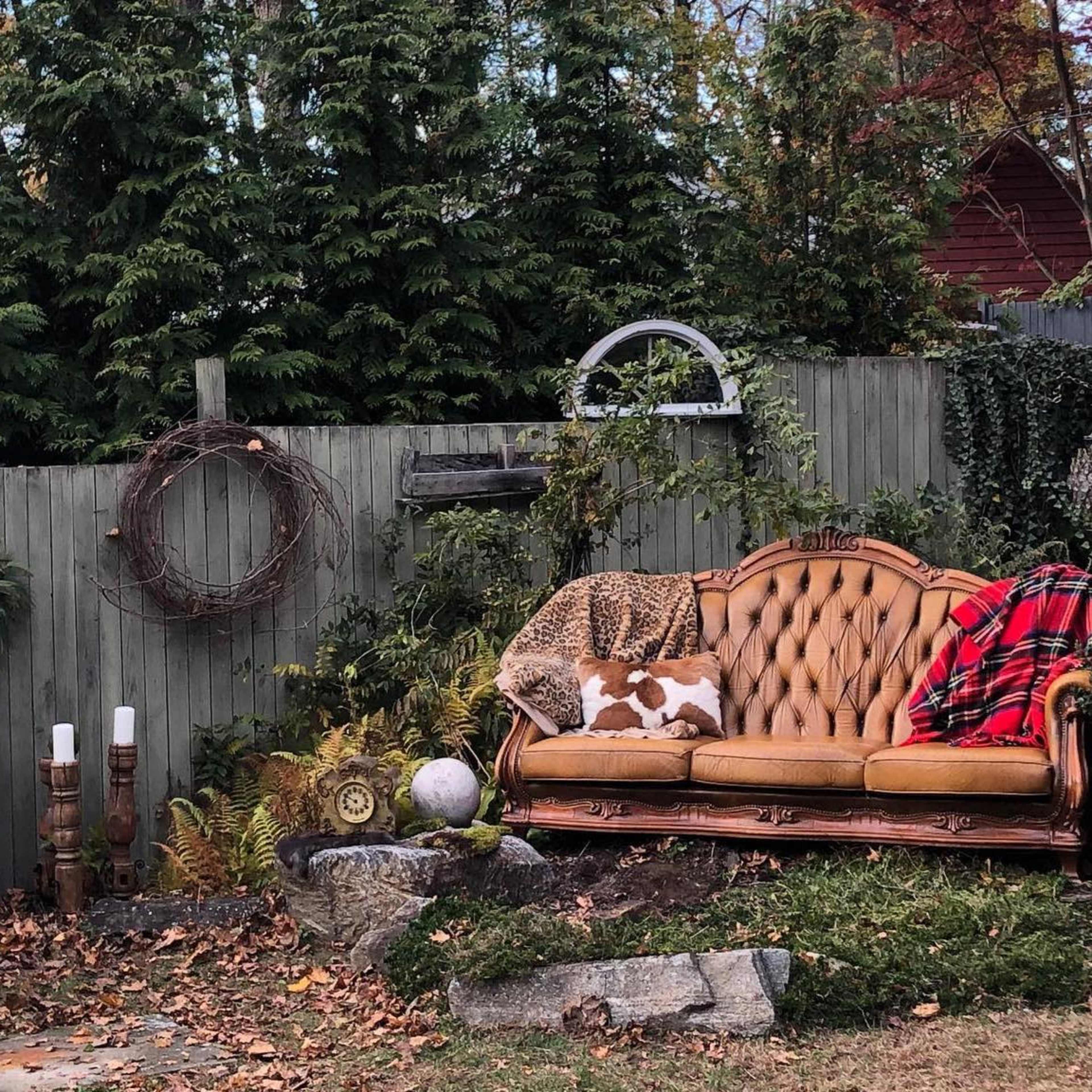 An outdoor seating area with a brown velvet sofa, a matching armchair, and various decorative elements against a wooden fence.