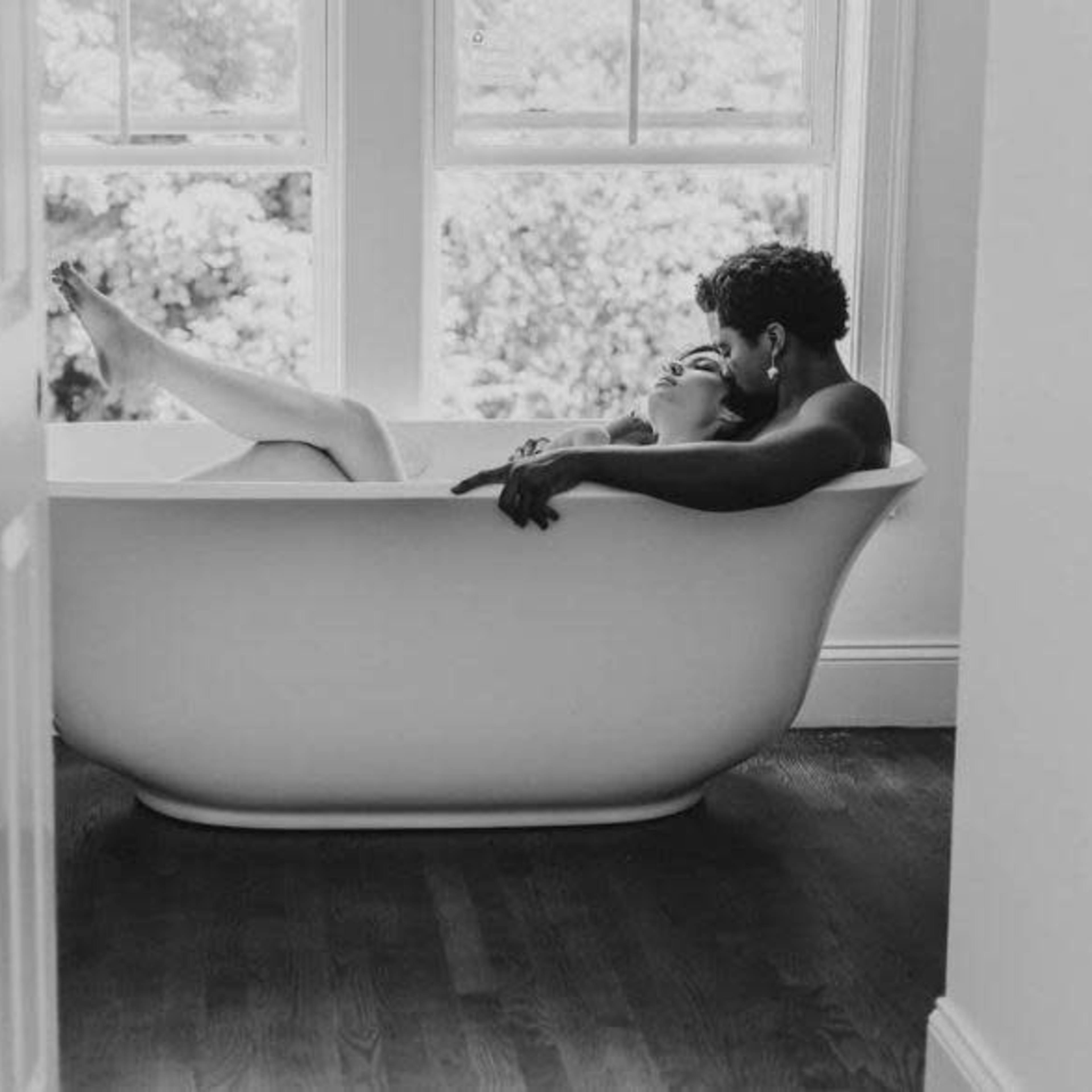 A man and a woman are taking a bath together in a large white tub near a window.
