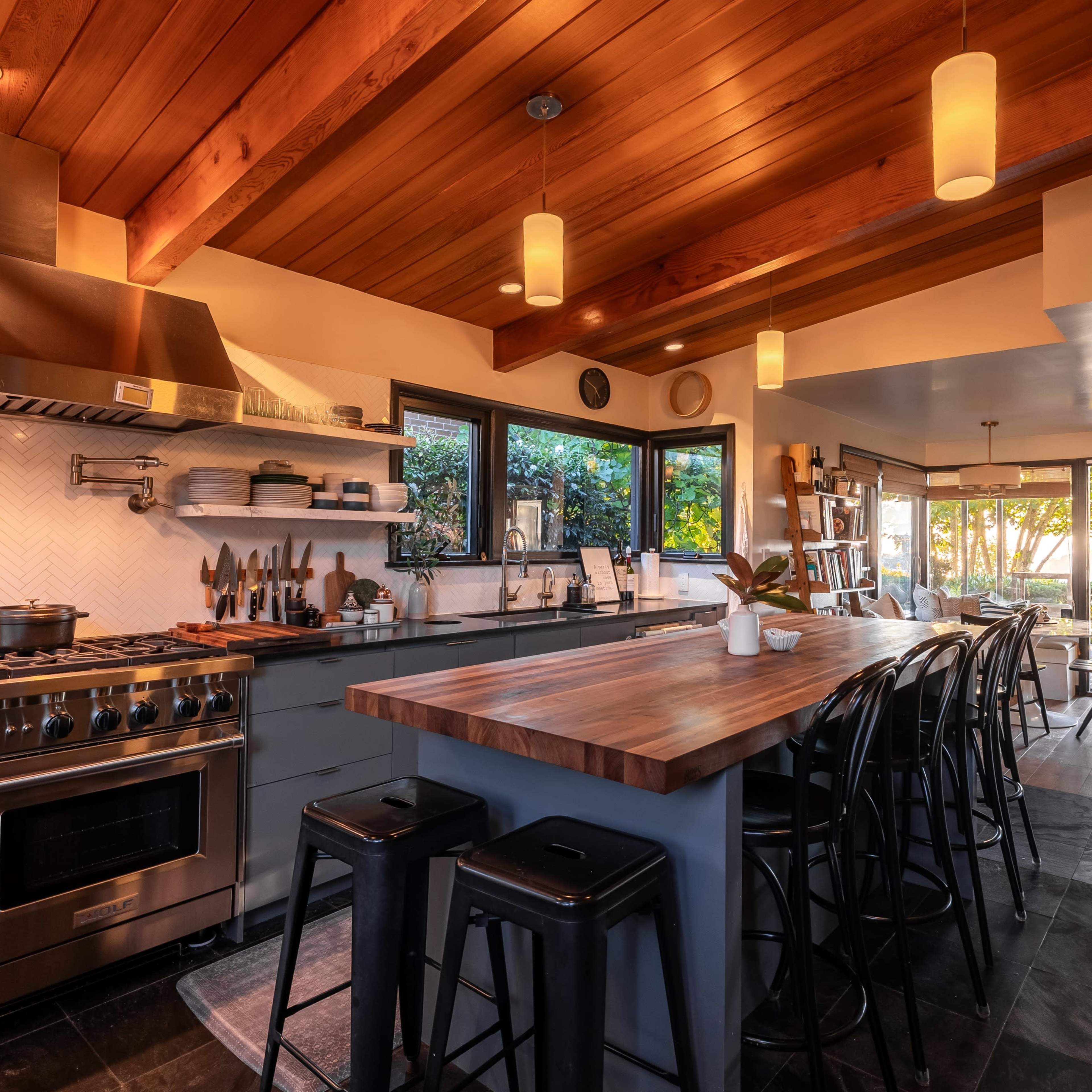 The modern kitchen features a large wooden island with bar seating, stainless steel appliances, and large windows overlooking a patio area.