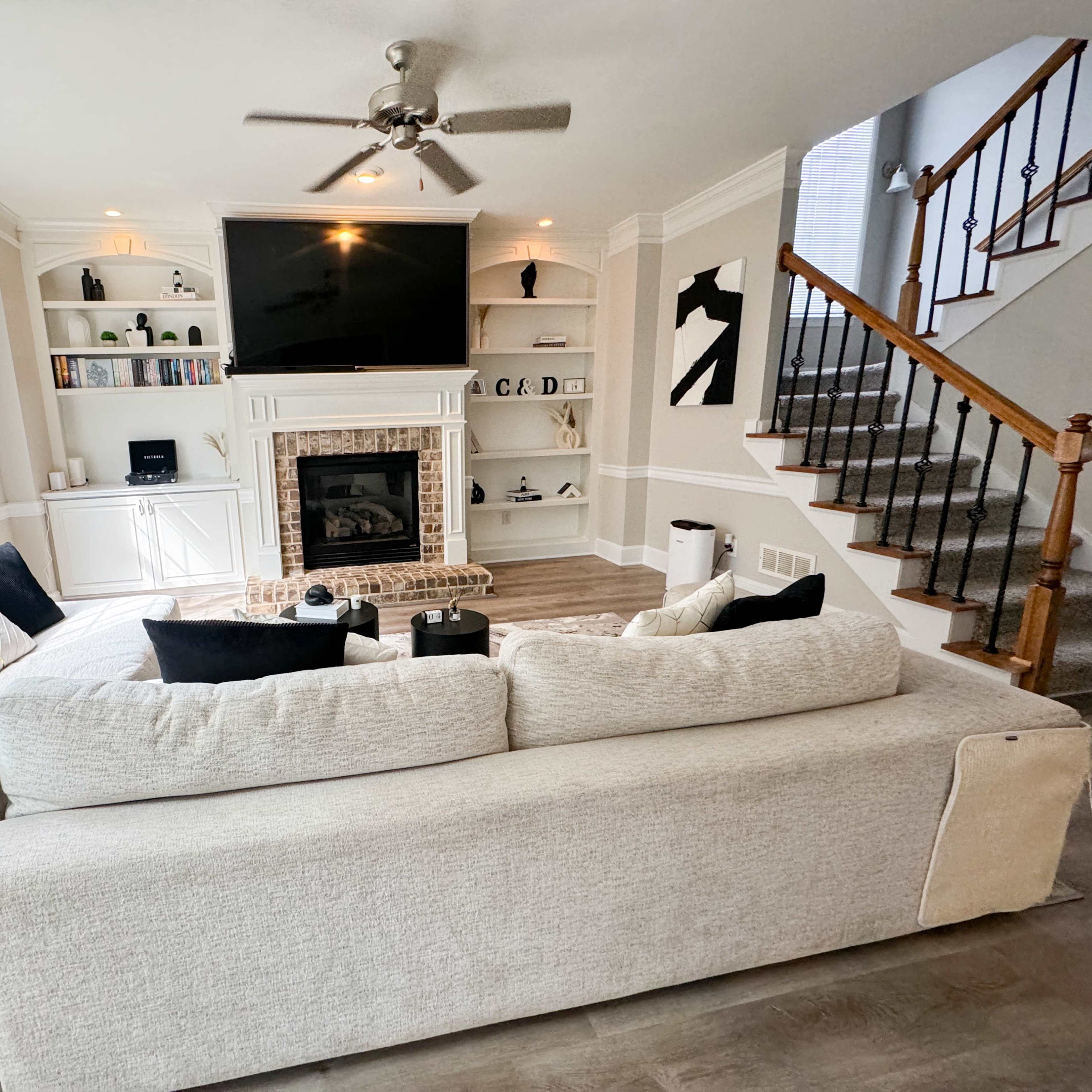 A spacious living room features a large sectional sofa, a brick fireplace, built-in shelving with a television above, and a staircase leading to an upper level.