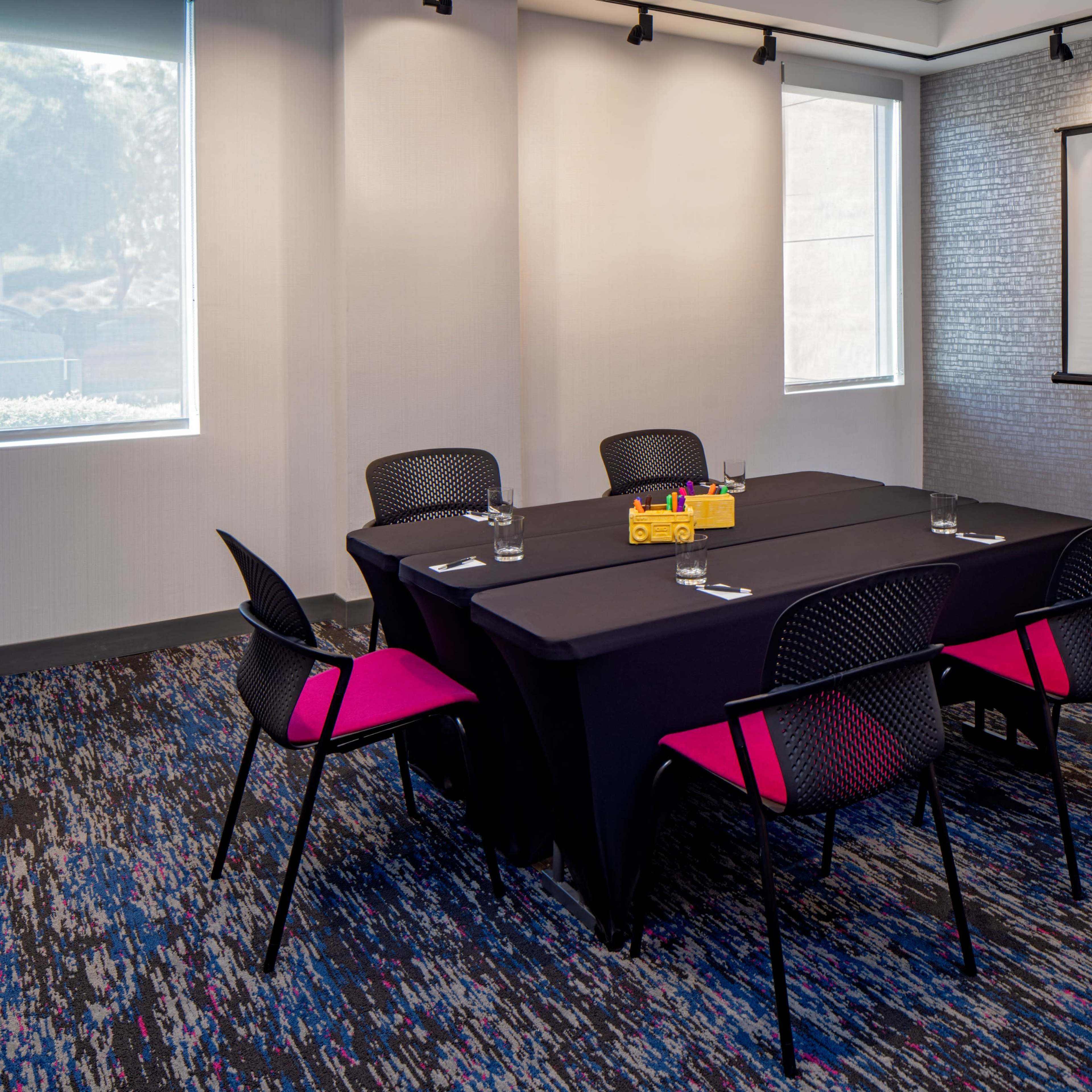 A small conference room features a black table with five chairs, a projector screen on a stand, and a colorful rug.