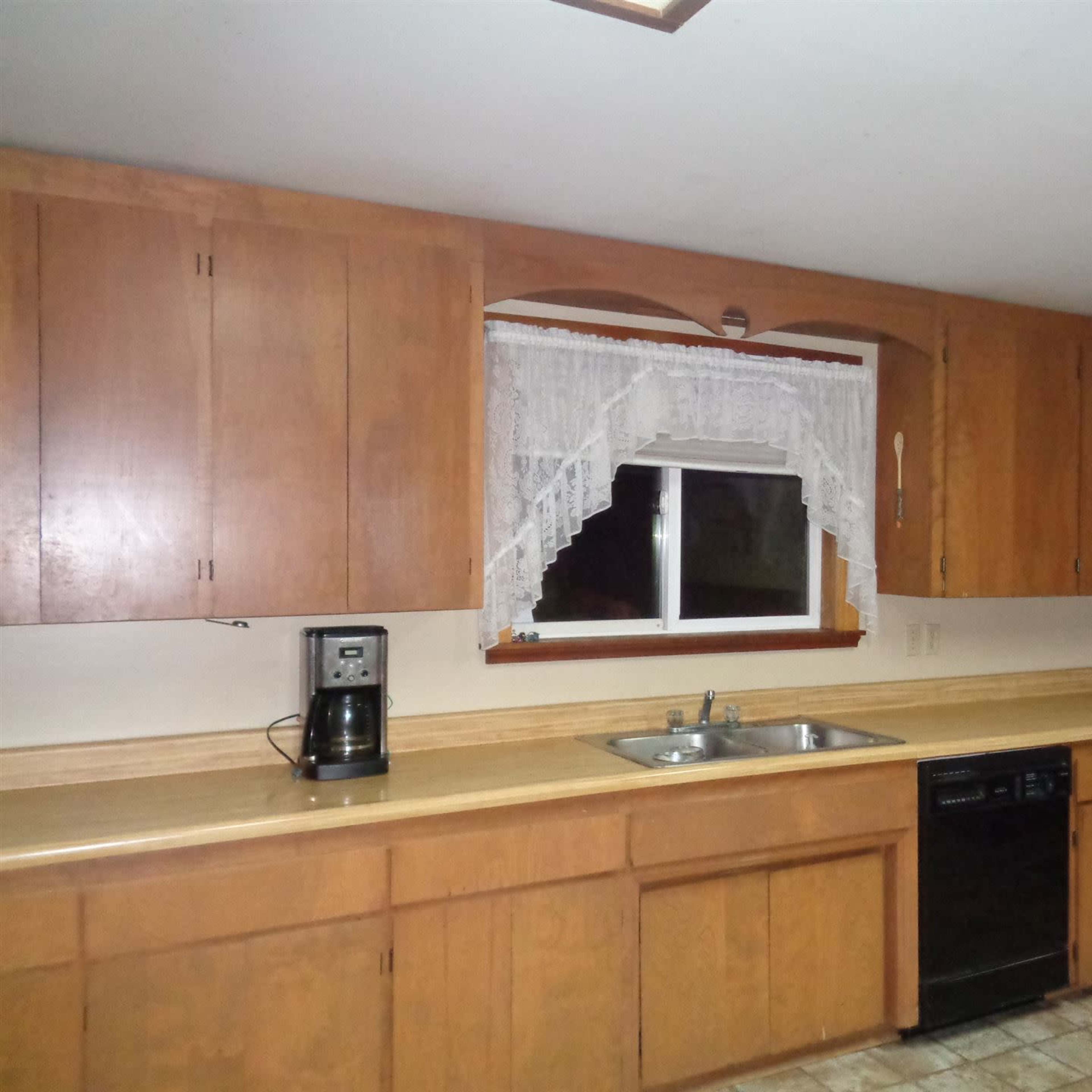 The kitchen features wooden cabinets, a window with lace curtains, a double sink, a coffee maker, a dishwasher, and a refrigerator along the counters.
