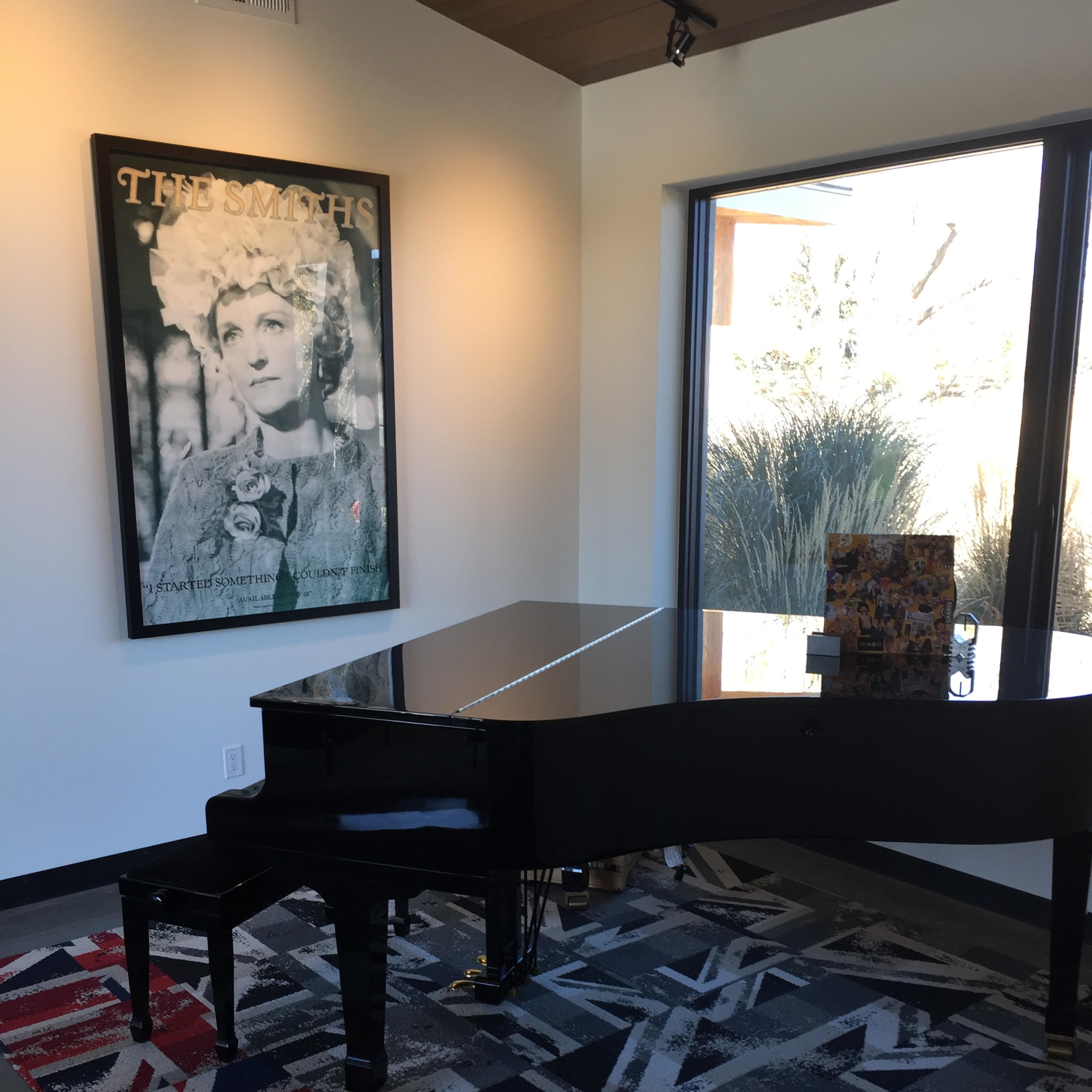 A black grand piano sits on a patterned rug in front of a window, with a framed poster of a woman on the wall.