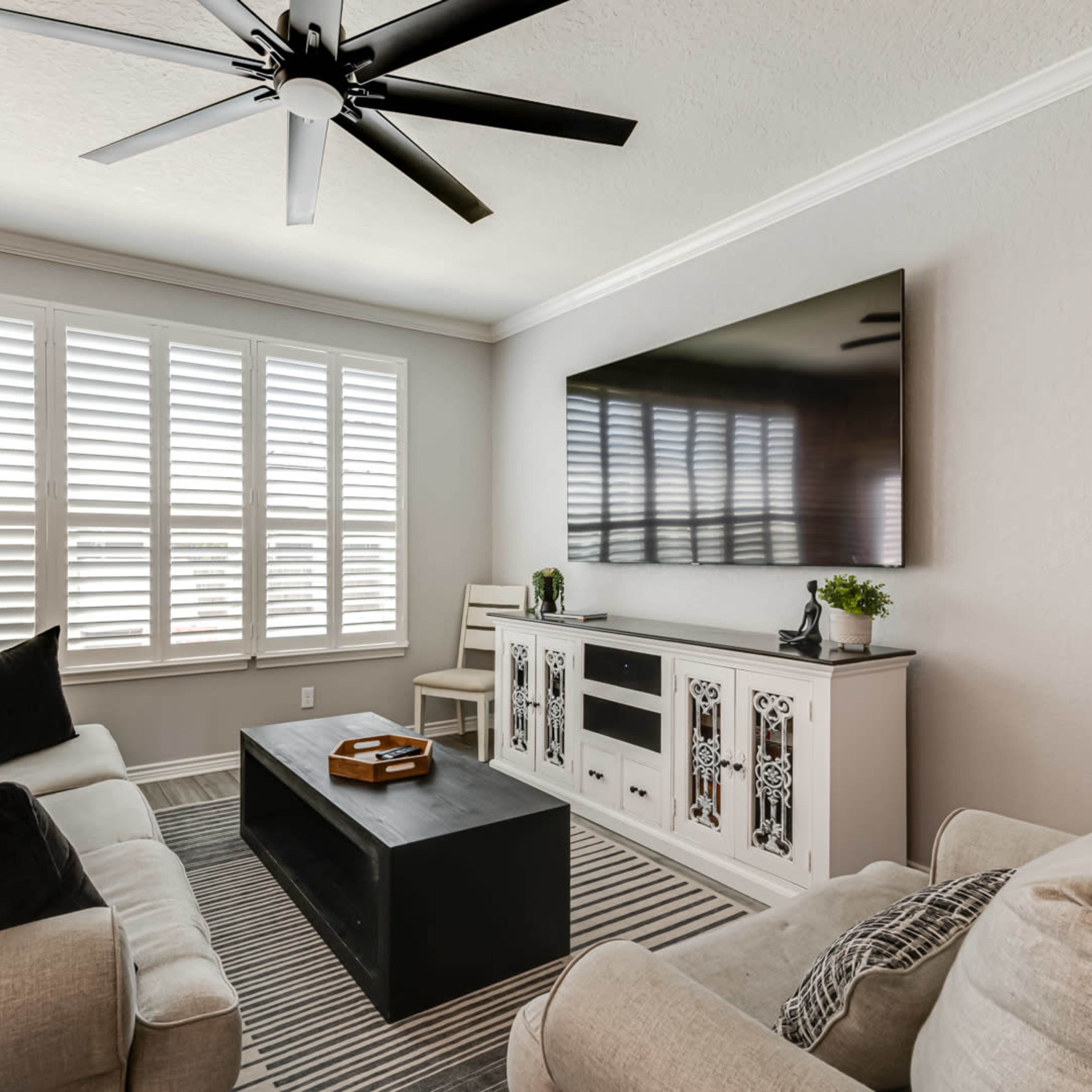 A living room features two sofas, a coffee table, a television mounted on the wall, and shuttered windows.