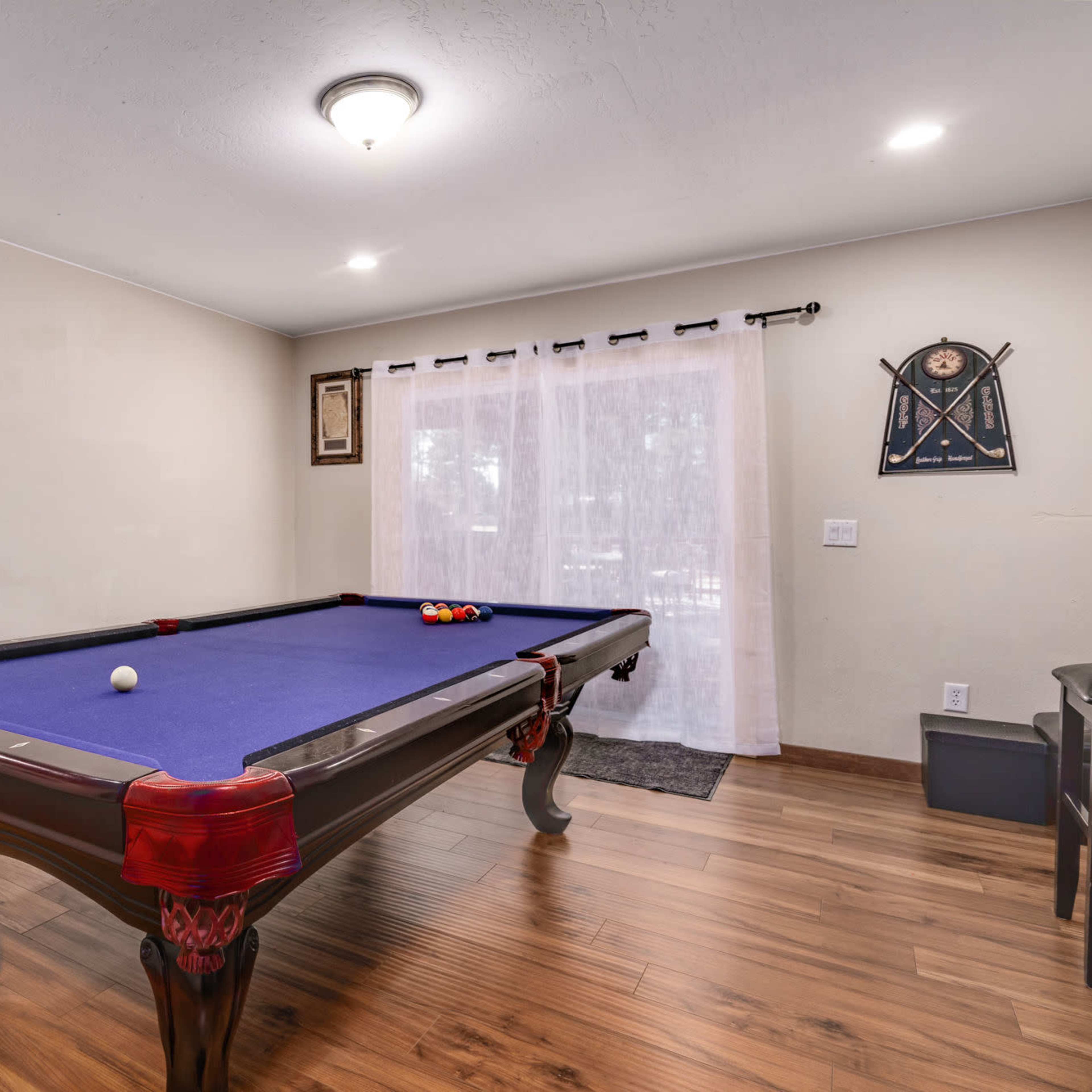 A pool table with balls arranged on it placed in a room with wooden flooring and walls adorned with framed decorations.