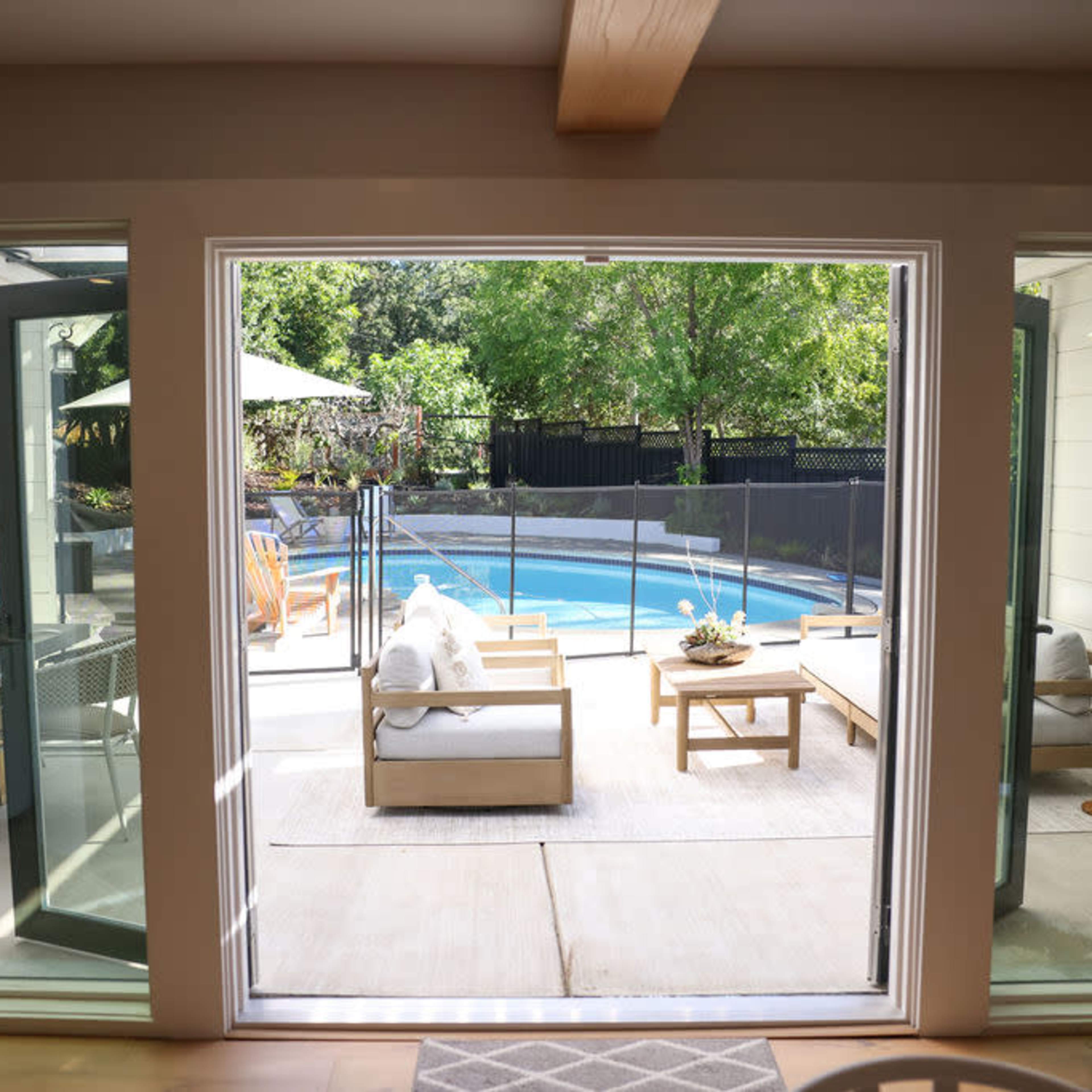 A modern living space with large windows opens up to a patio and a swimming pool surrounded by greenery.