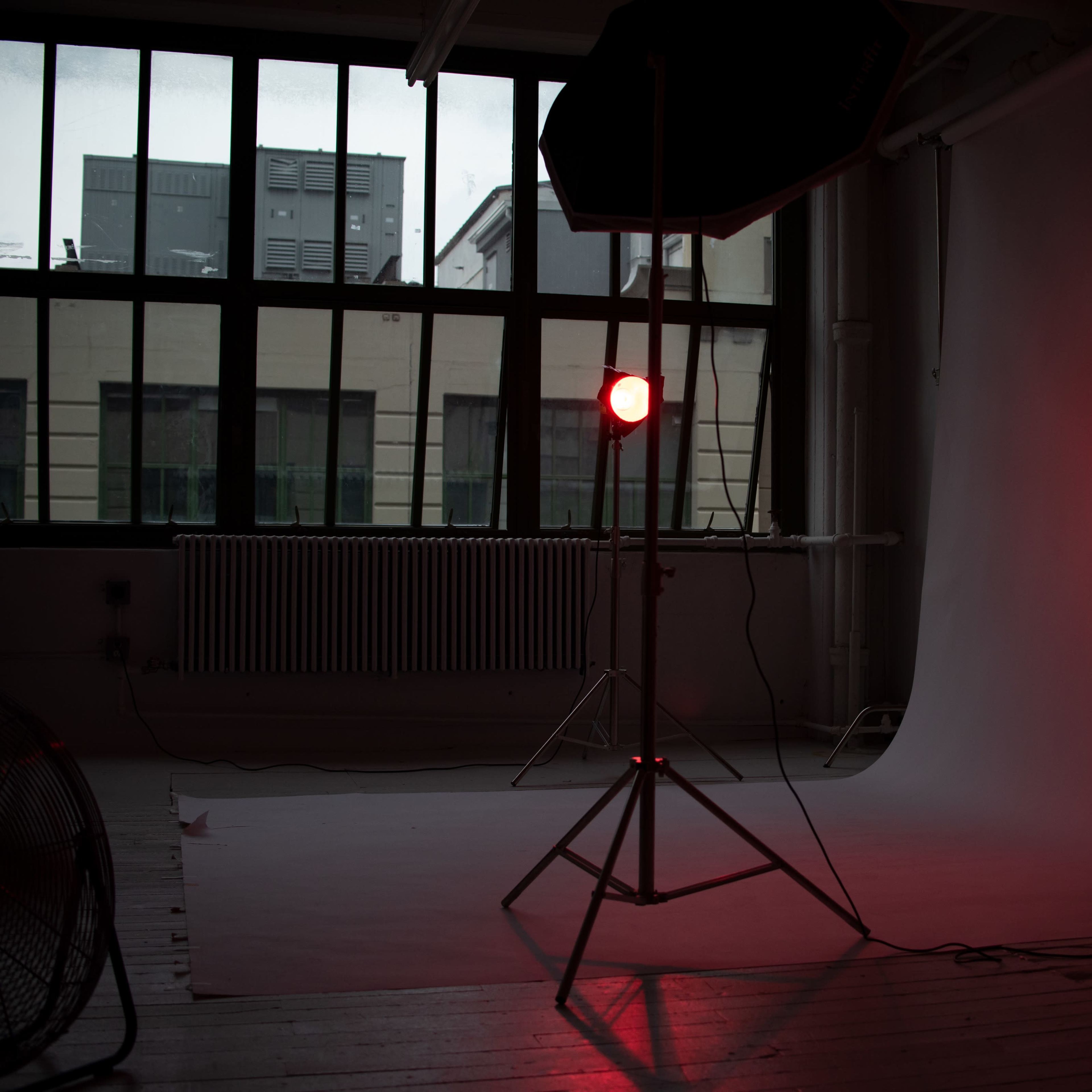 A photography studio is shown with a red light mounted on a stand, positioned next to a white backdrop and large windows.