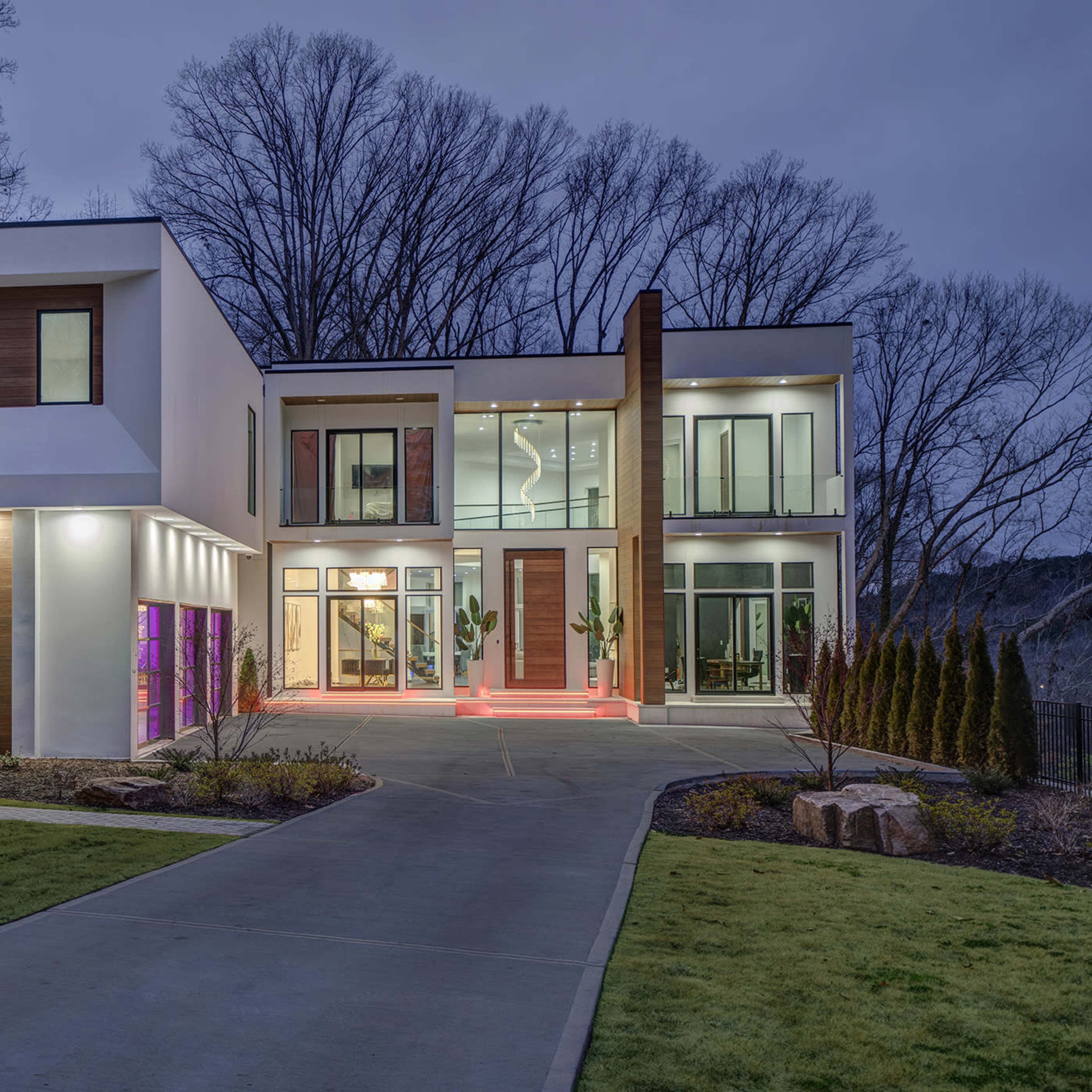 A modern two-story house with large glass windows and a landscaped yard is illuminated in the twilight.