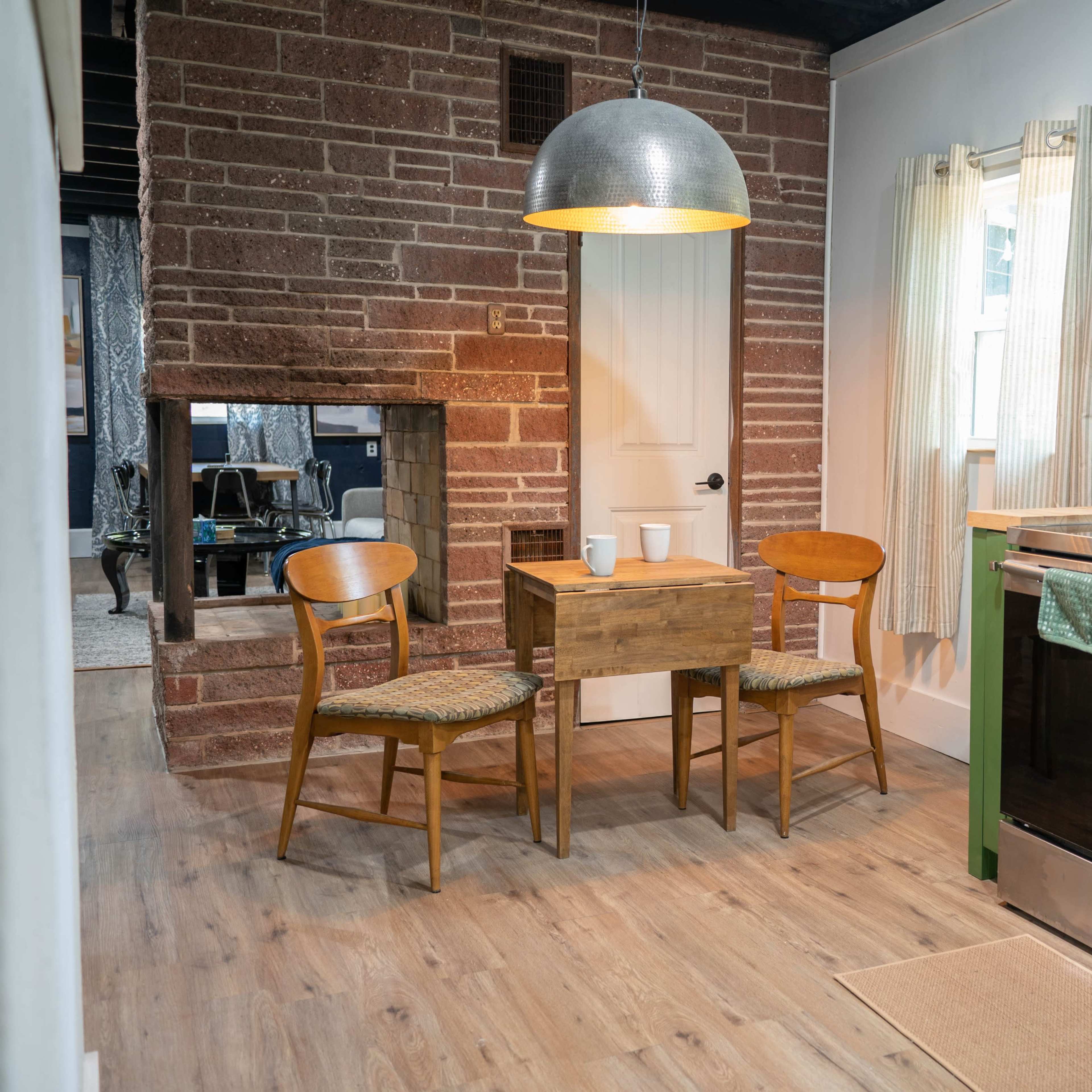 A small kitchen features a wooden table and two chairs, a green cabinetry setup, a stainless steel stove, and a rustic brick wall.