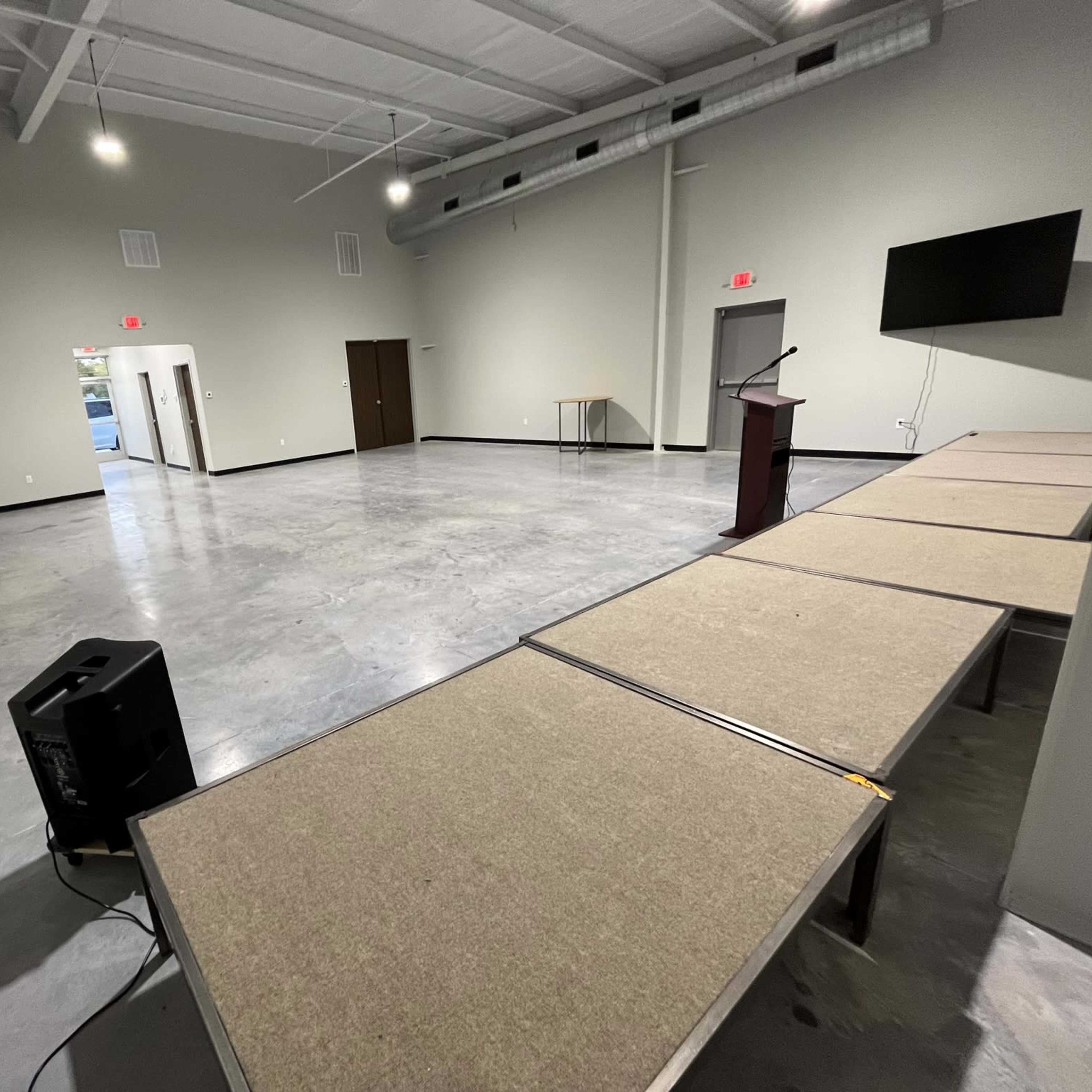 A spacious, empty room with gray floors, tables arranged in a line, and a podium facing a wall-mounted screen.