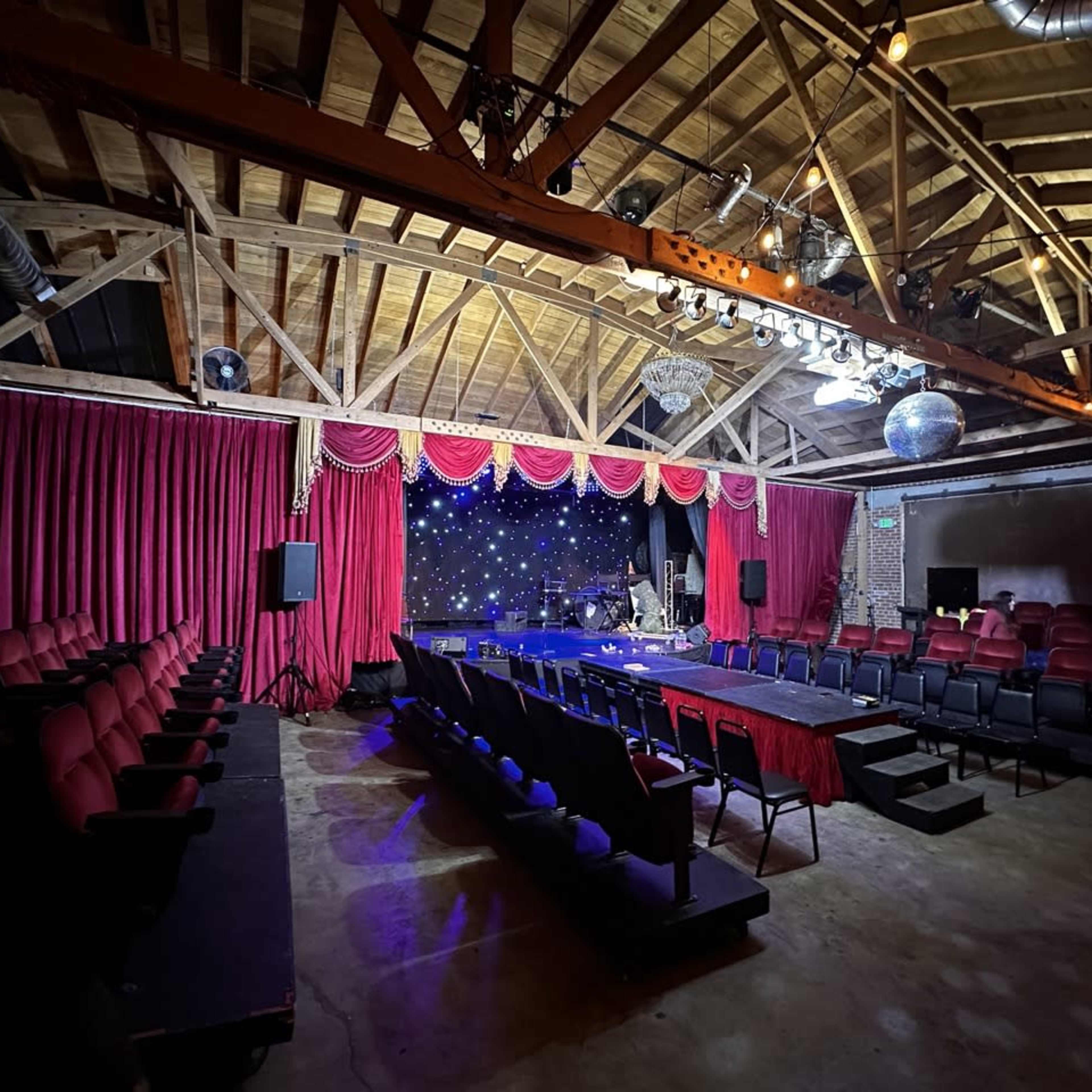 An empty theater with red velvet curtains, a stage adorned with twinkling lights, and rows of red and black seats arranged for an audience.