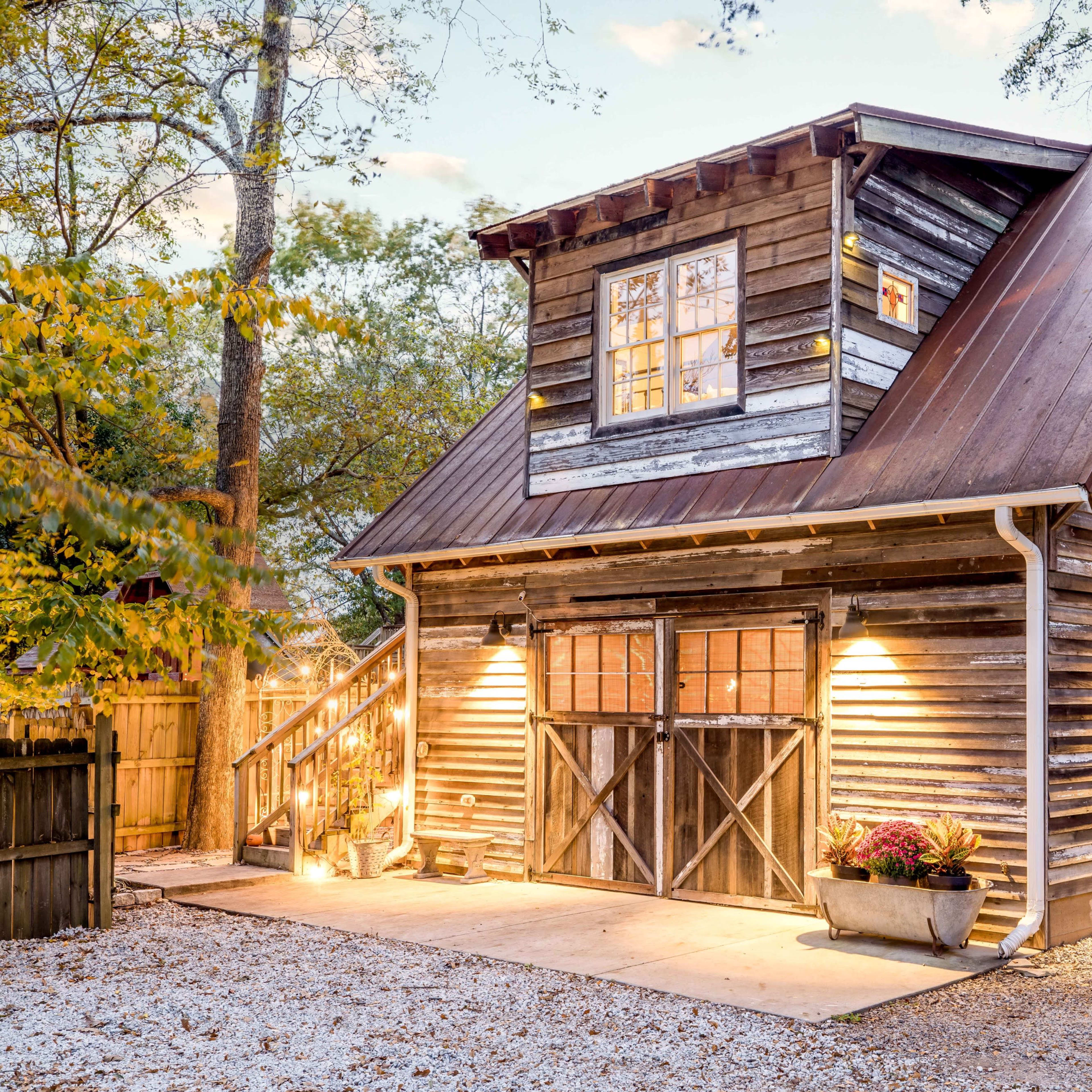 A rustic wooden house with a metal roof, illuminated by soft outdoor lighting, surrounded by trees and a wooden fence.