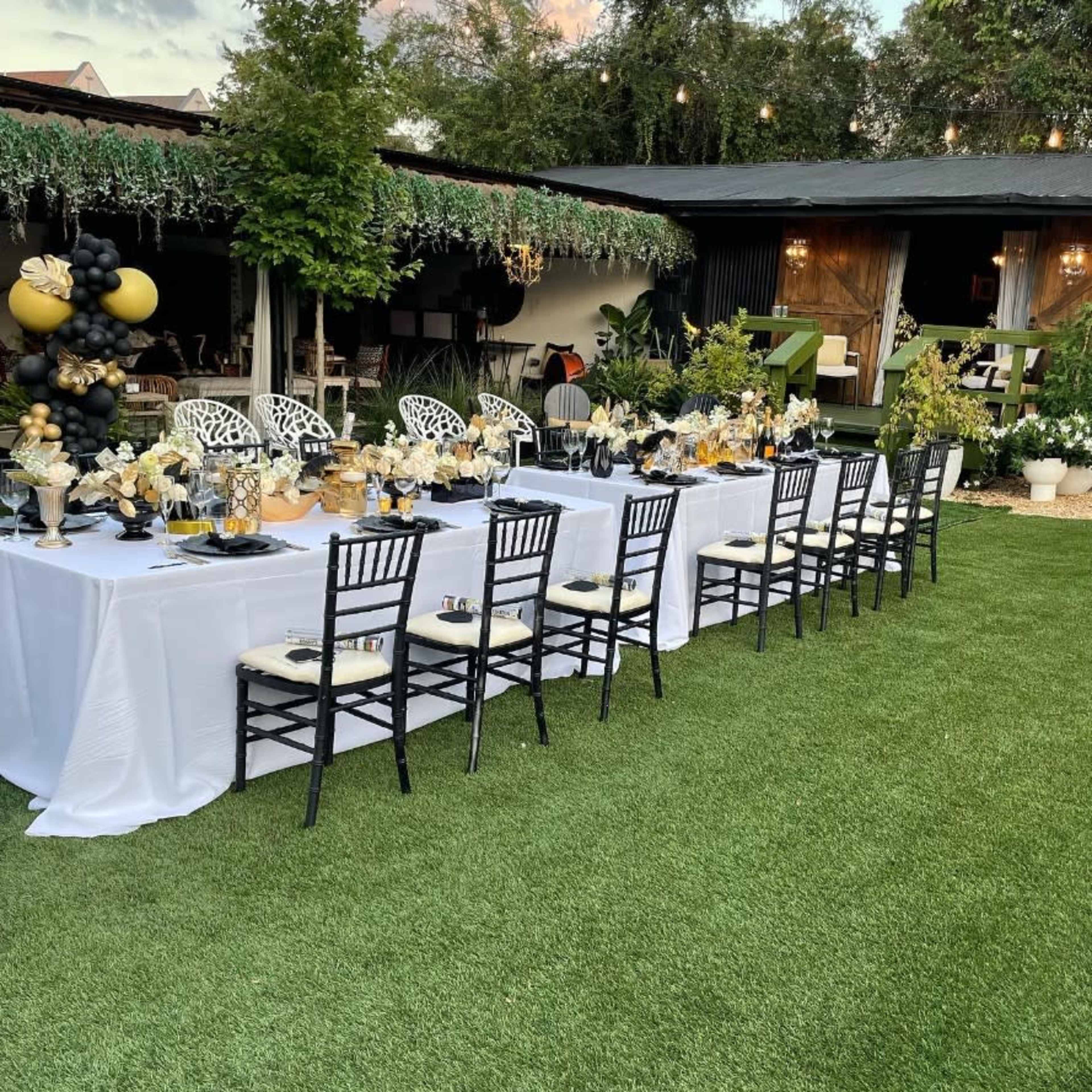 A long banquet table is set outdoors with elegant decor, surrounded by greenery and string lights.