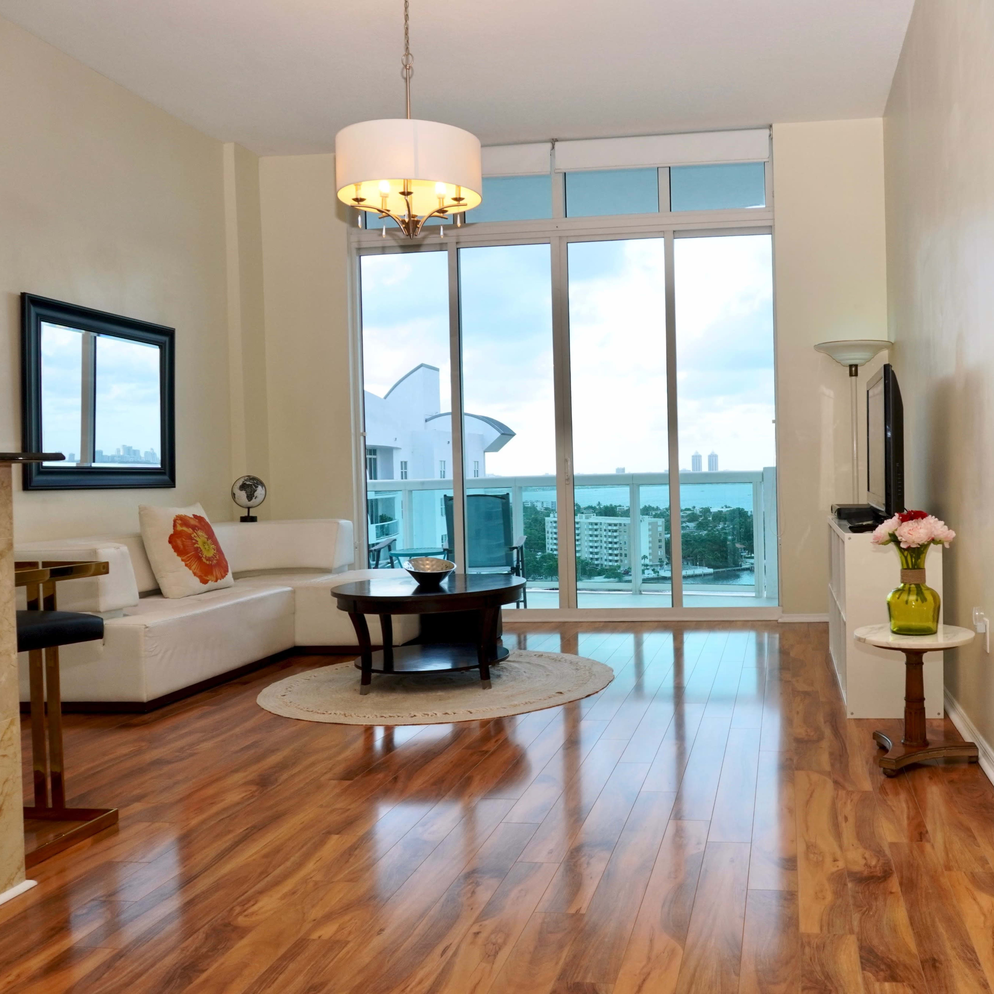 The scene shows a modern living room with wooden flooring, large windows providing a view, and a minimalist seating area featuring a white couch and a round rug.