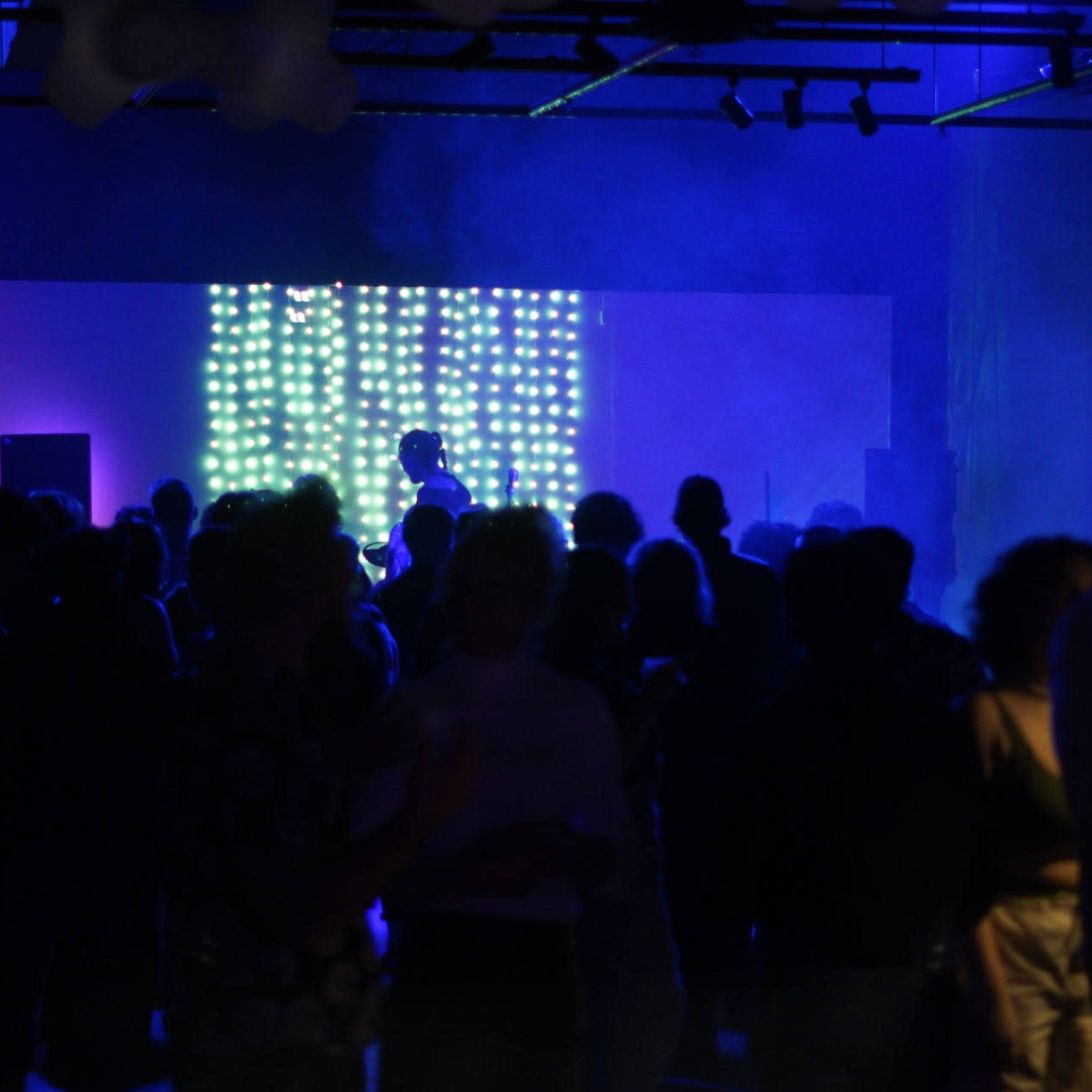 A crowd of people dances in a dimly lit venue, illuminated by a backdrop of colorful lights.
