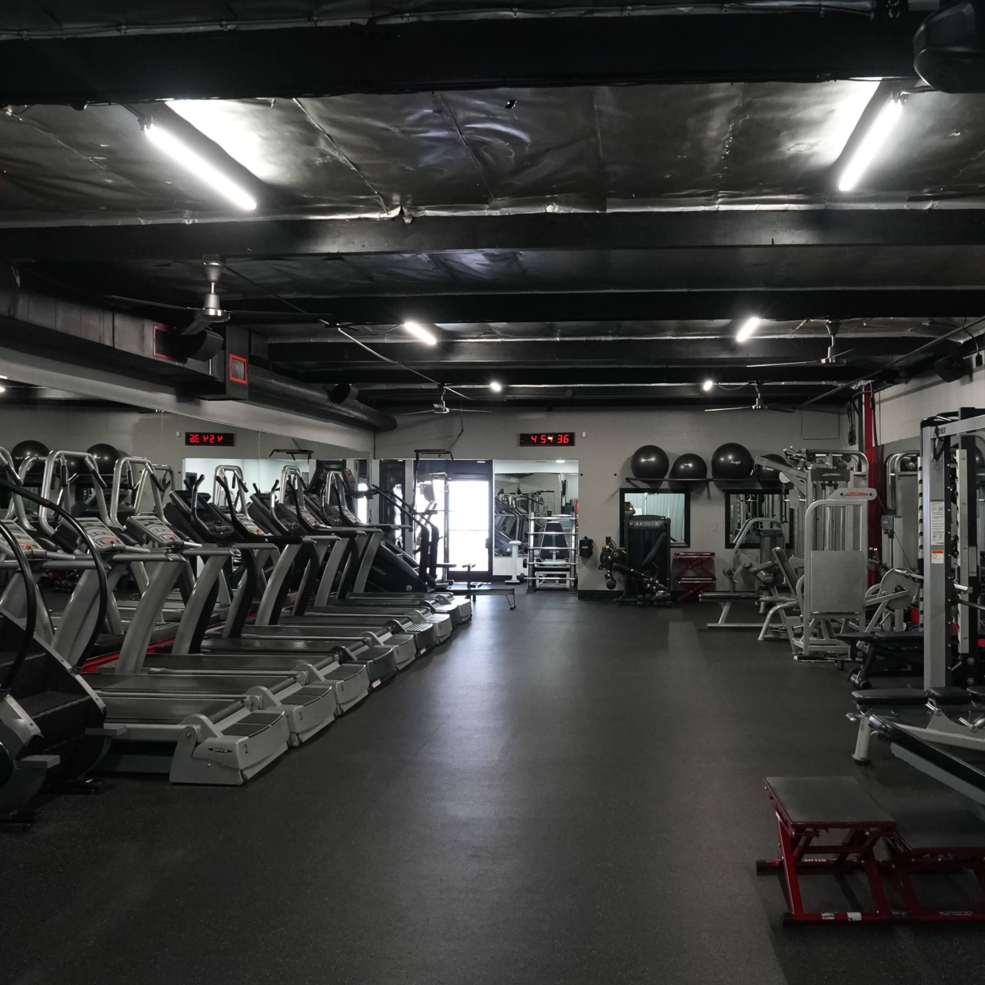 A modern gym interior with various exercise machines arranged in rows, including treadmills and weight machines, illuminated by overhead lights.