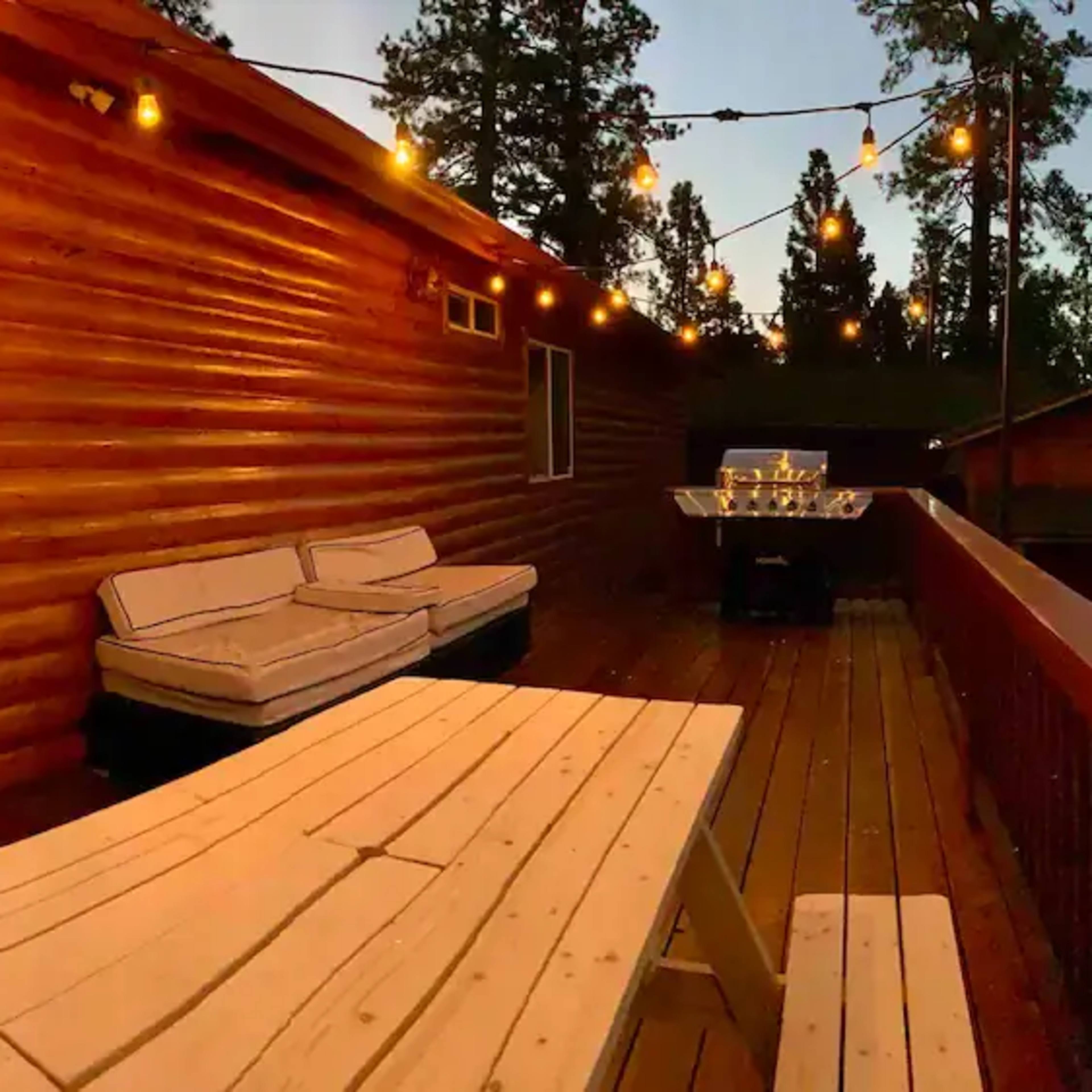 A wooden deck with a table and seating area, illuminated by string lights, next to a log cabin and a barbecue setup.