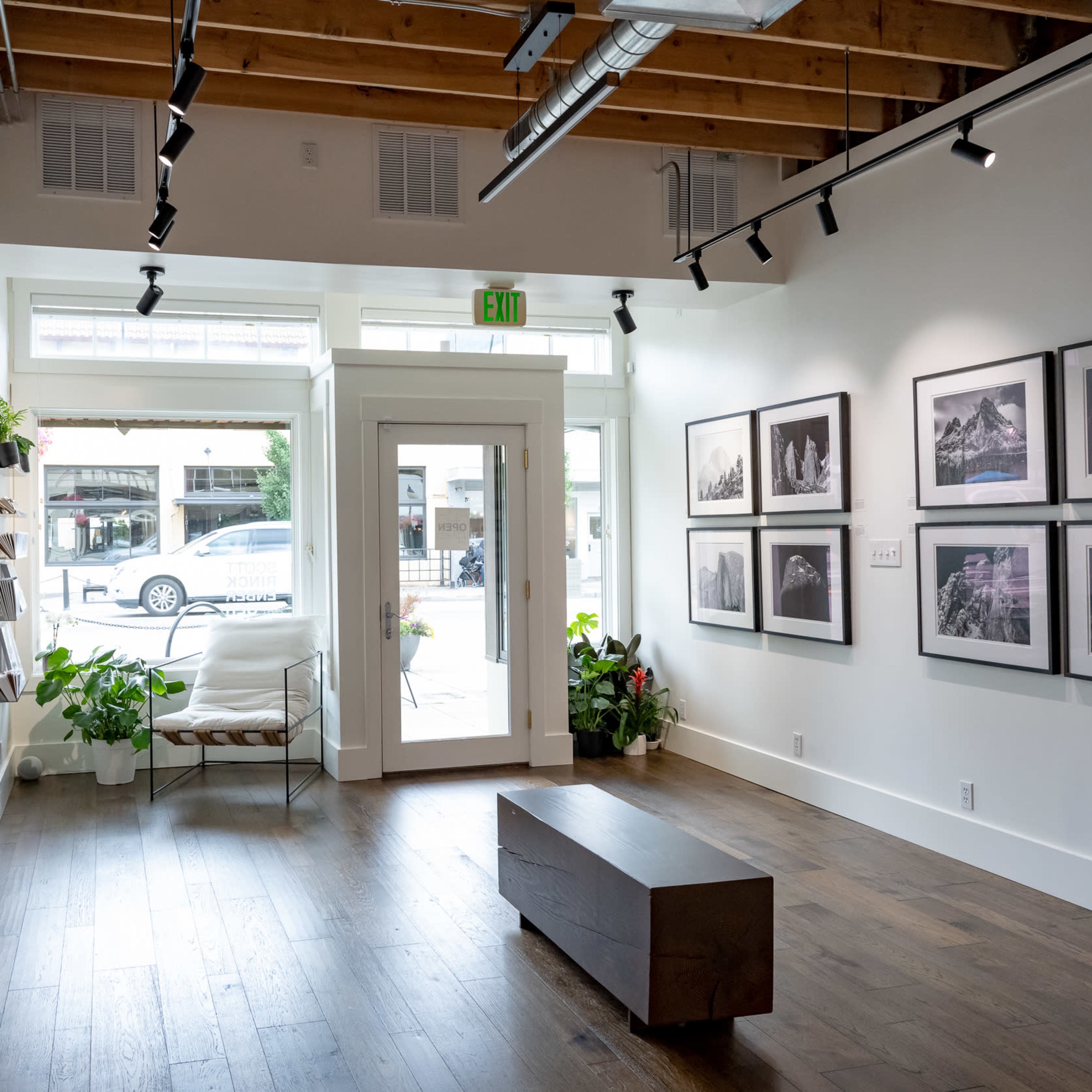 A gallery space with framed photographs on the walls, a wooden bench in the center, and large windows allowing natural light to enter.