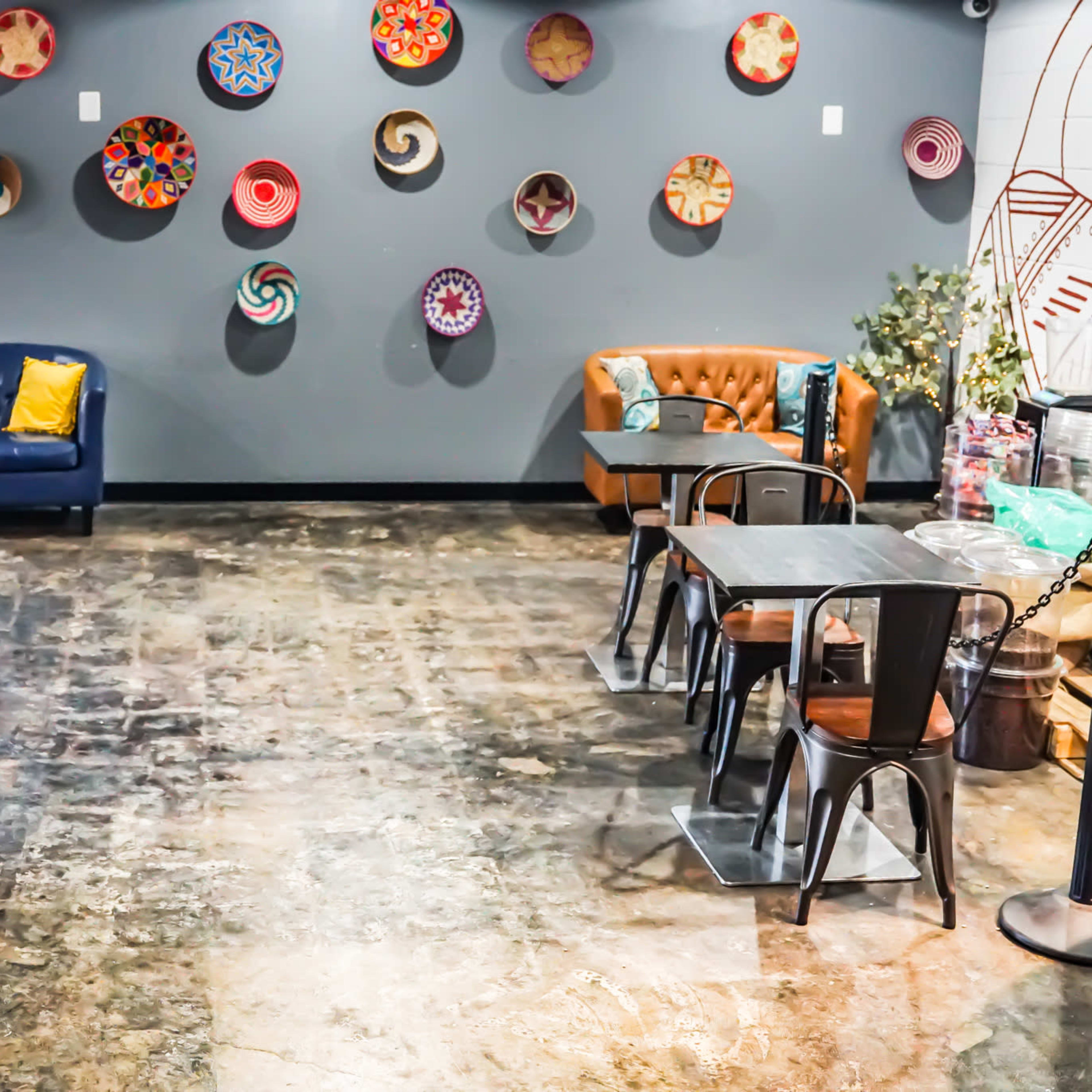 A cozy café interior with a blue sofa, brown leather seating, and tables arranged on a polished concrete floor, adorned with colorful wall decorations.