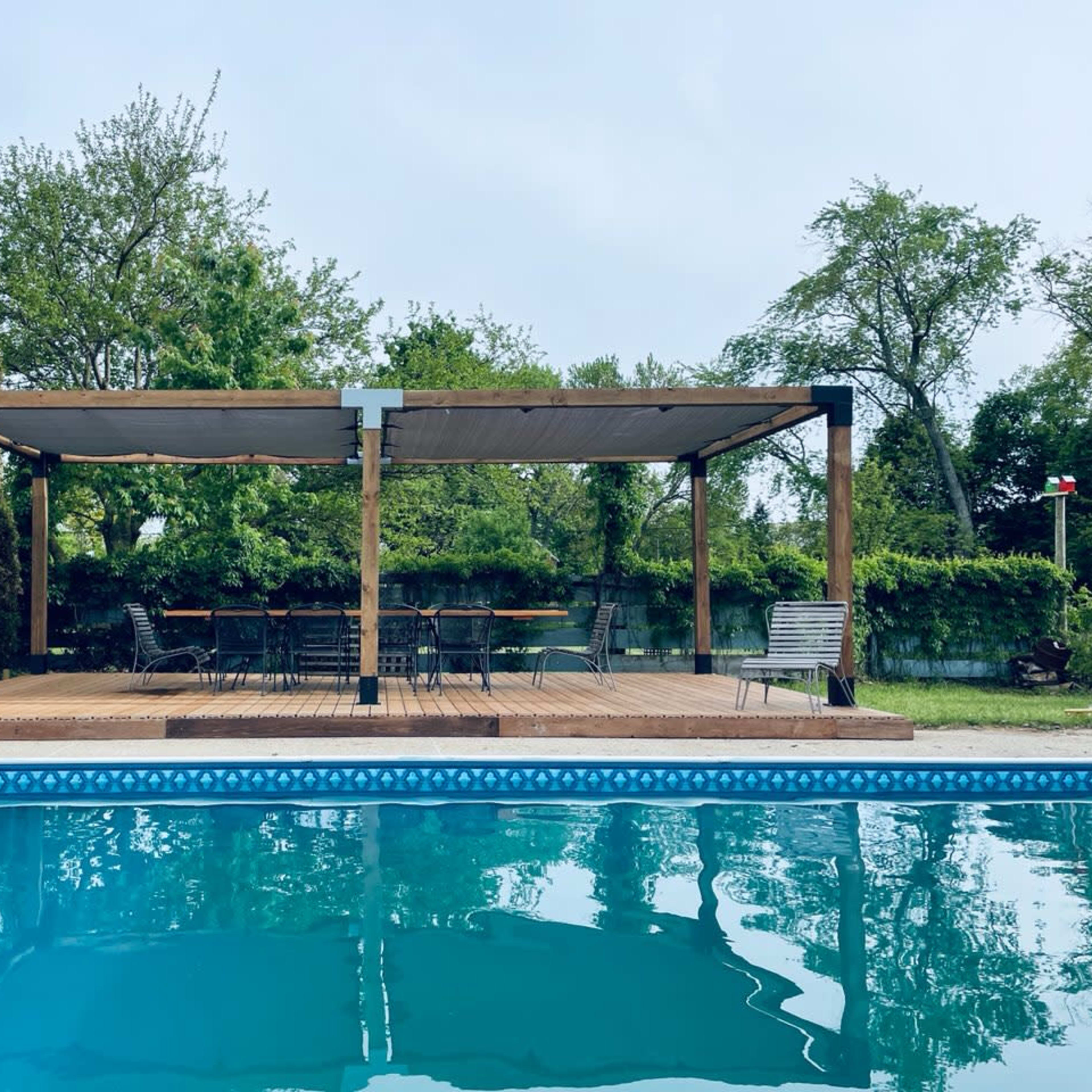 A wooden deck with a pergola overlooks a clear swimming pool surrounded by greenery.