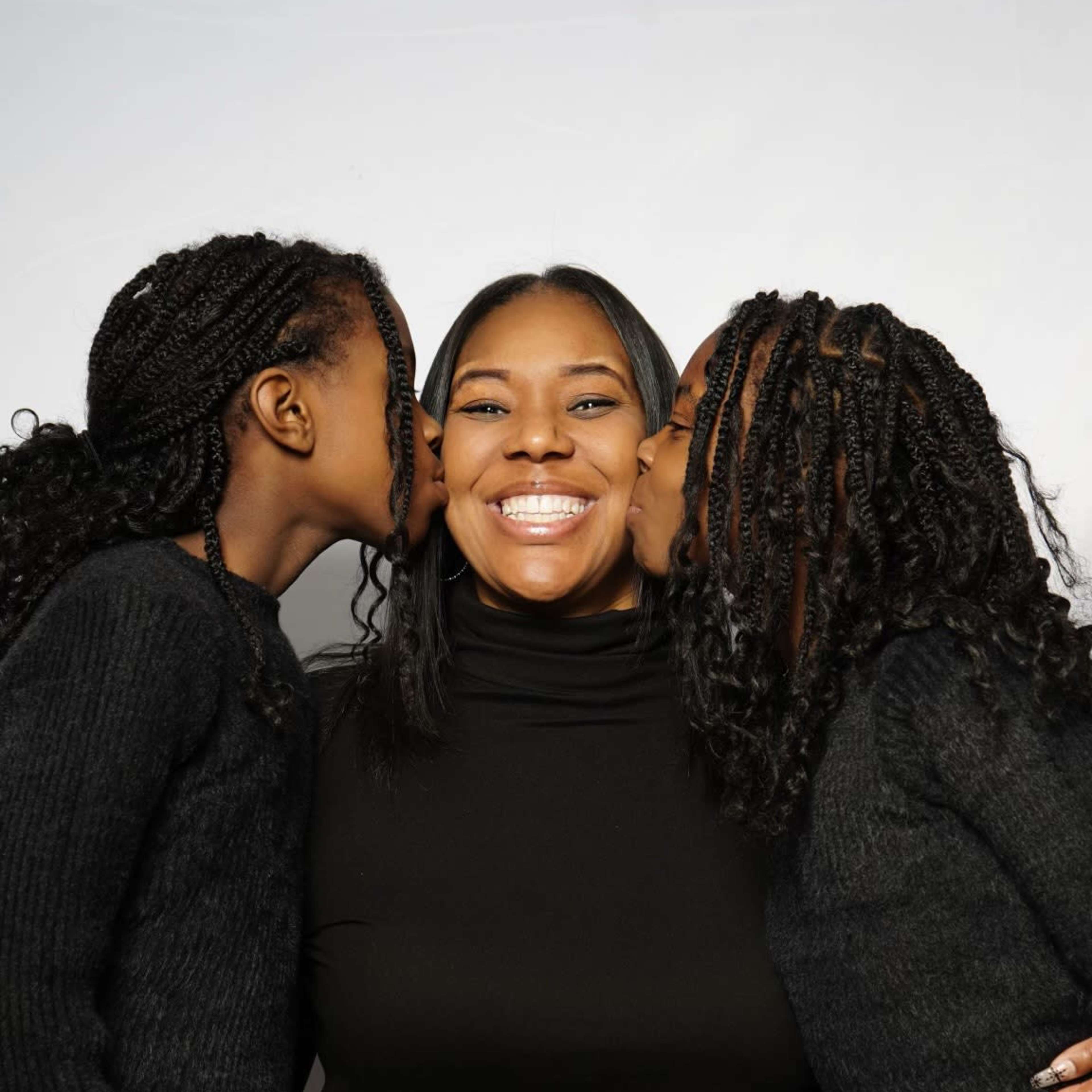 A woman sits smiling with two girls on either side, each giving her a kiss on the cheek.