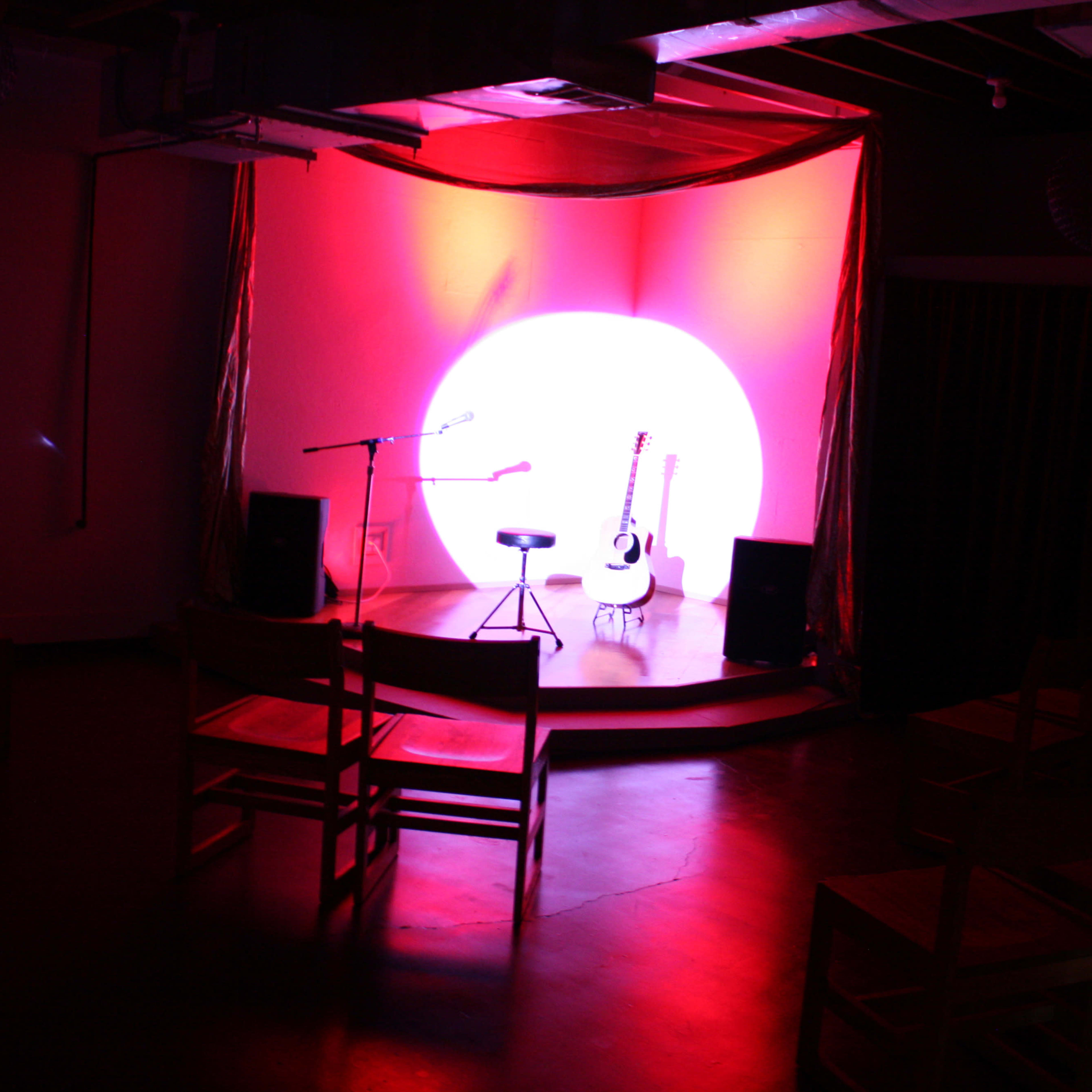 A spotlight illuminates an empty stage with a microphone, a stool, and an acoustic guitar in front of a large circular backdrop.