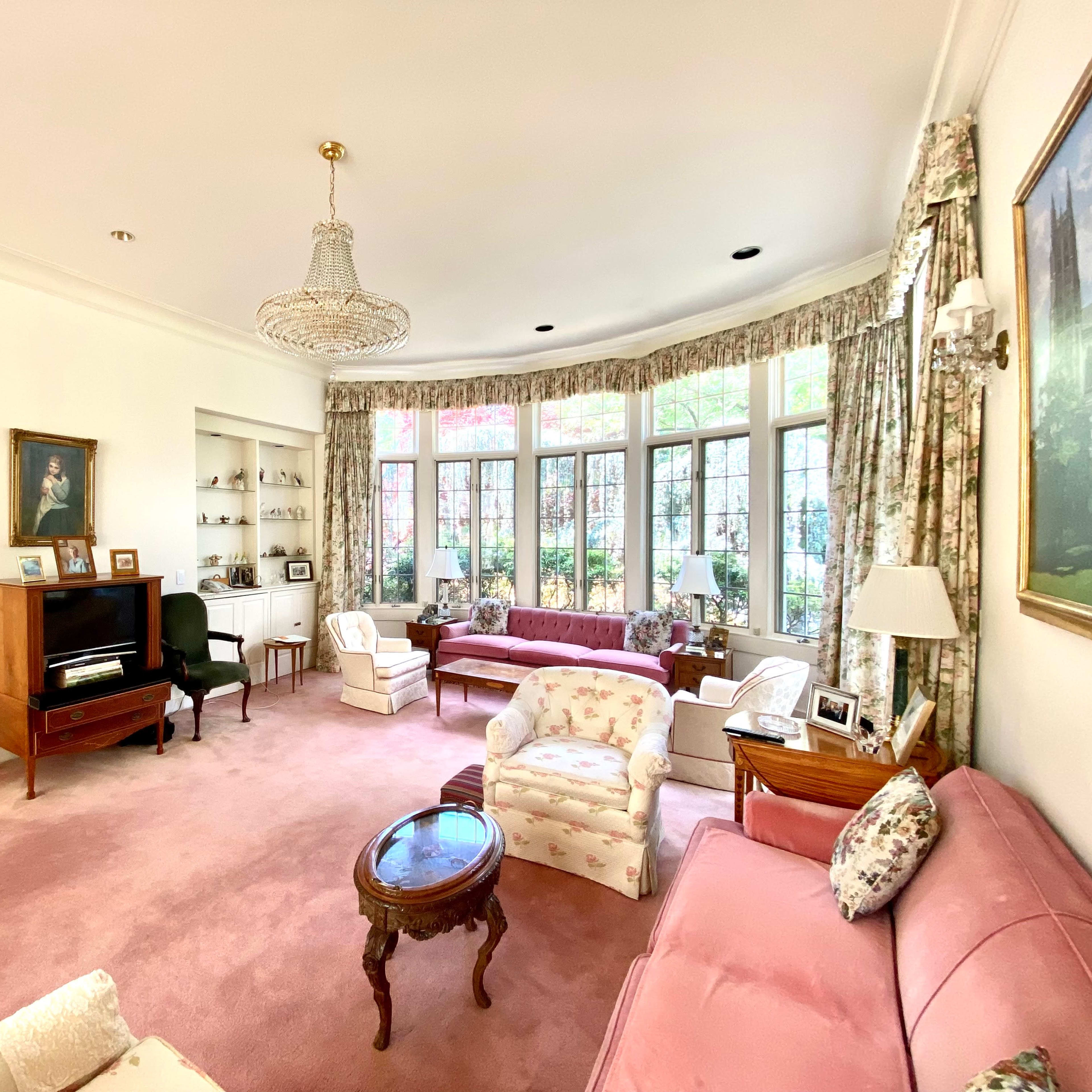The living room features a pink carpet, floral-patterned furniture, large windows with views of greenery, and a chandelier.