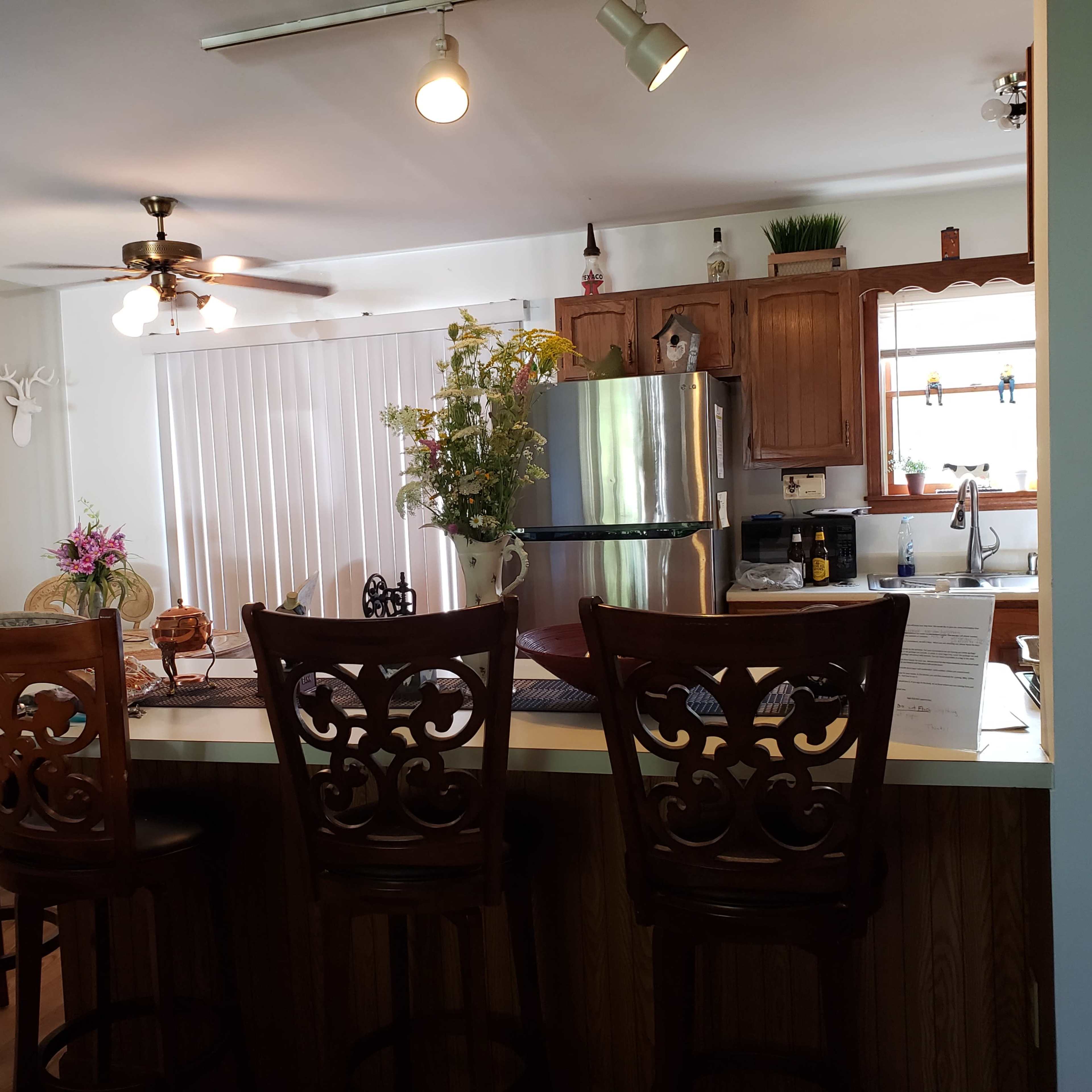 A kitchen features a bar with high stools, a stainless steel refrigerator, and a window overlooking a light-filled area with decorative plants and a deer-themed wall art.