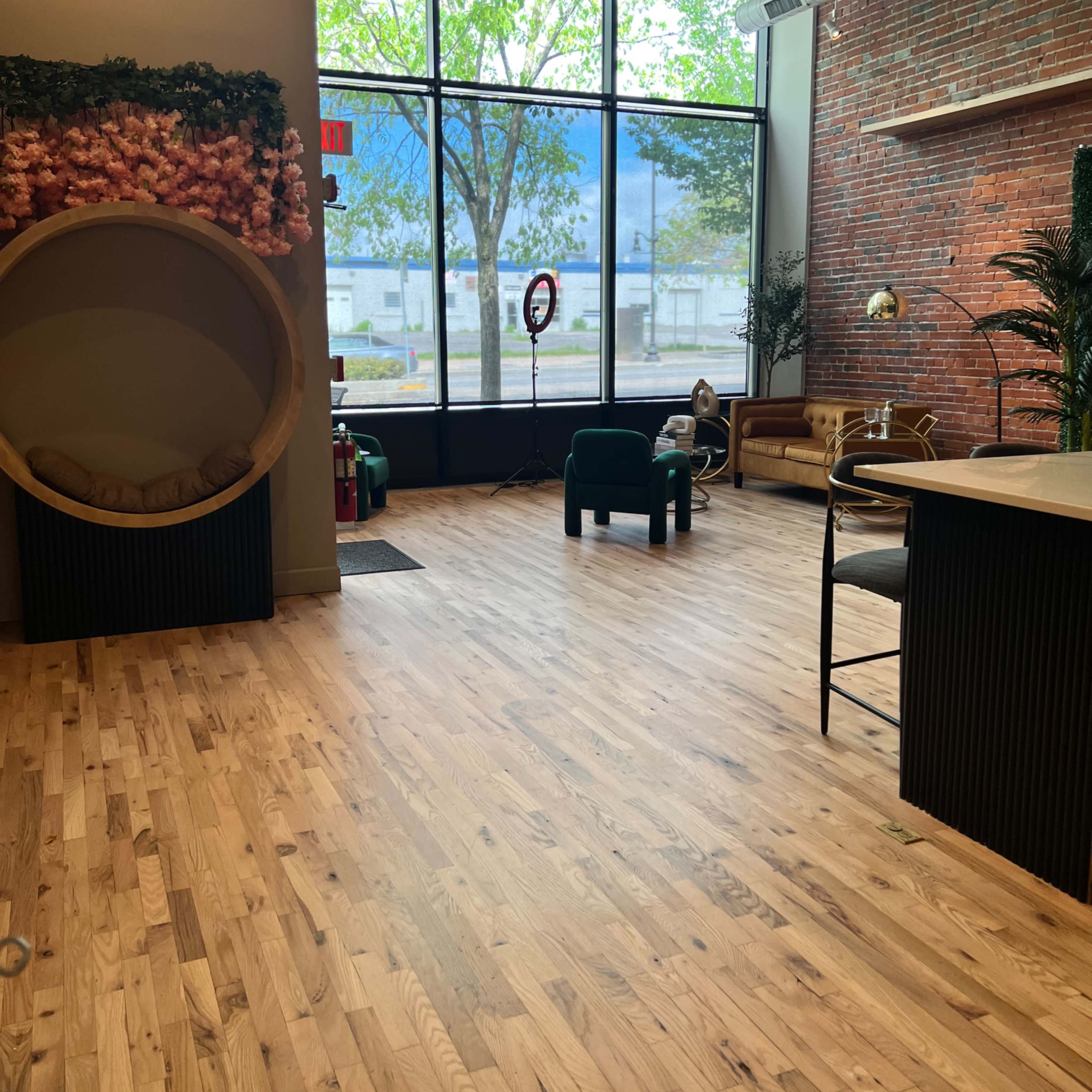 A spacious interior with wooden flooring, large windows letting in natural light, and a mix of furniture including a circular seating area, chairs, and decorative plants against a brick wall.