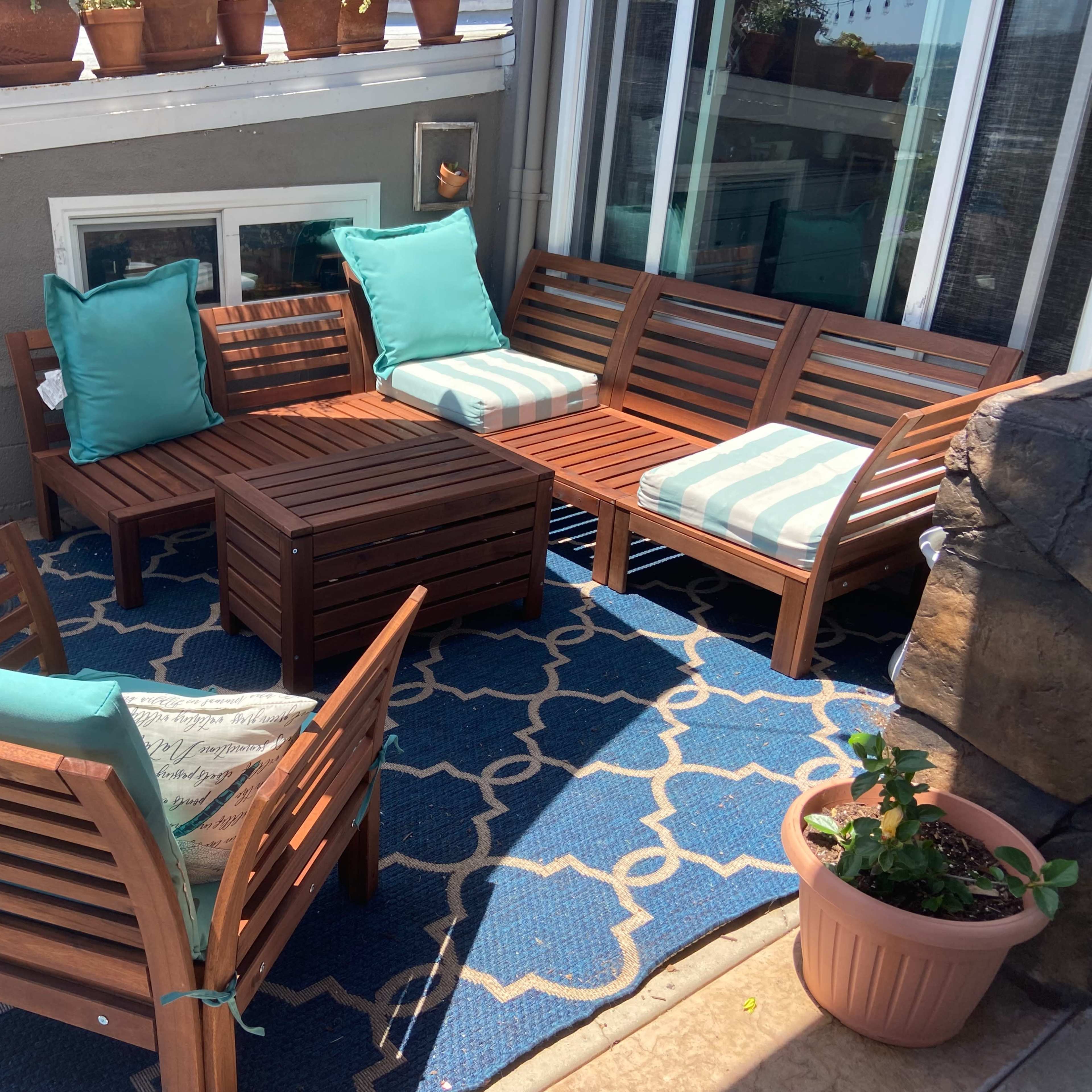 A wooden patio seating arrangement with turquoise cushions is placed on a blue patterned rug surrounded by potted plants.