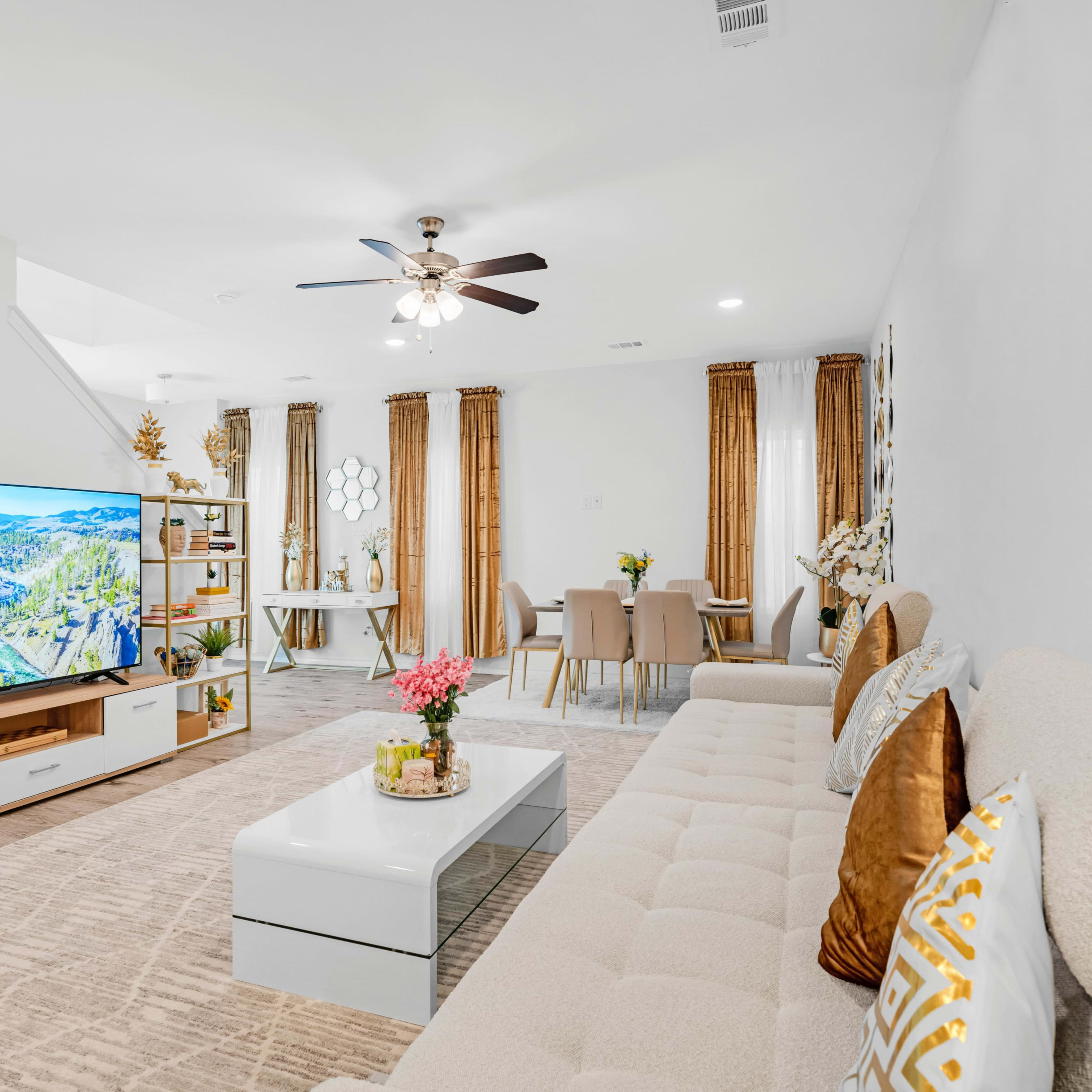 A modern living room features a large TV, a beige sectional sofa, a coffee table, and a dining area with a light-colored table set.