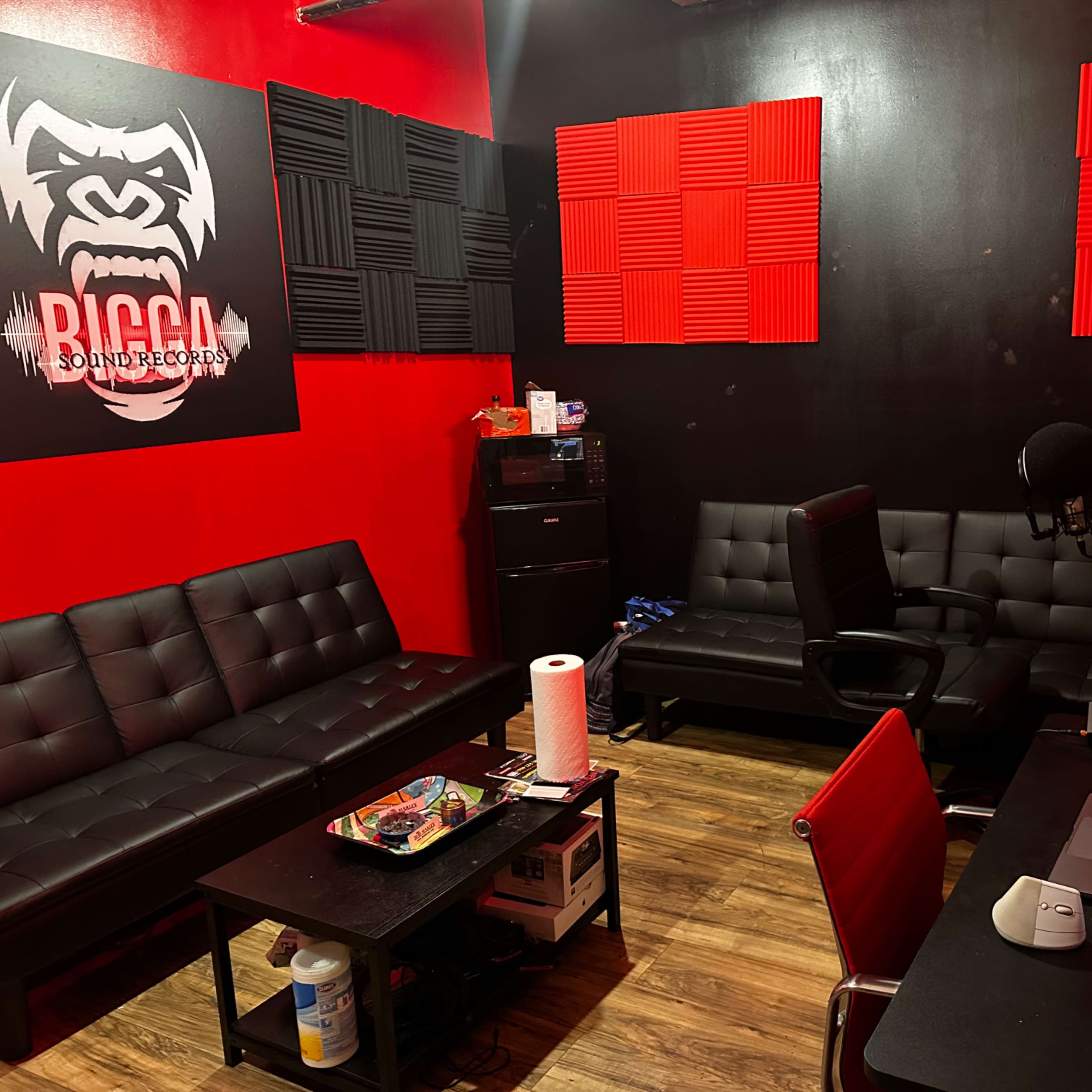 A music recording studio with black and red walls, featuring black couches, soundproof panels, a microphone, speakers, and a computer set up on a desk.