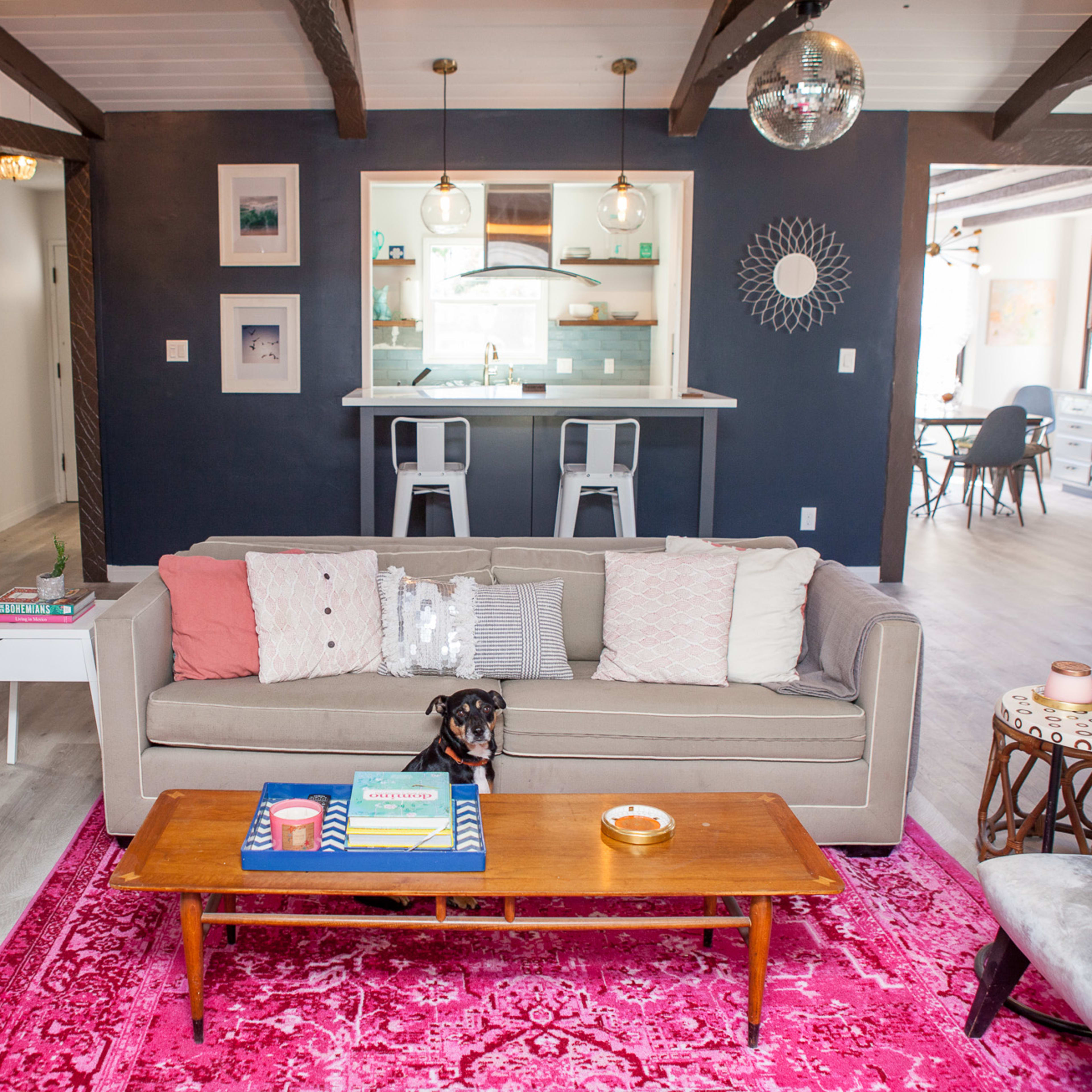 A modern living room features a beige couch with decorative pillows, a wooden coffee table, and a pink area rug, with a dining area and kitchen visible in the background.