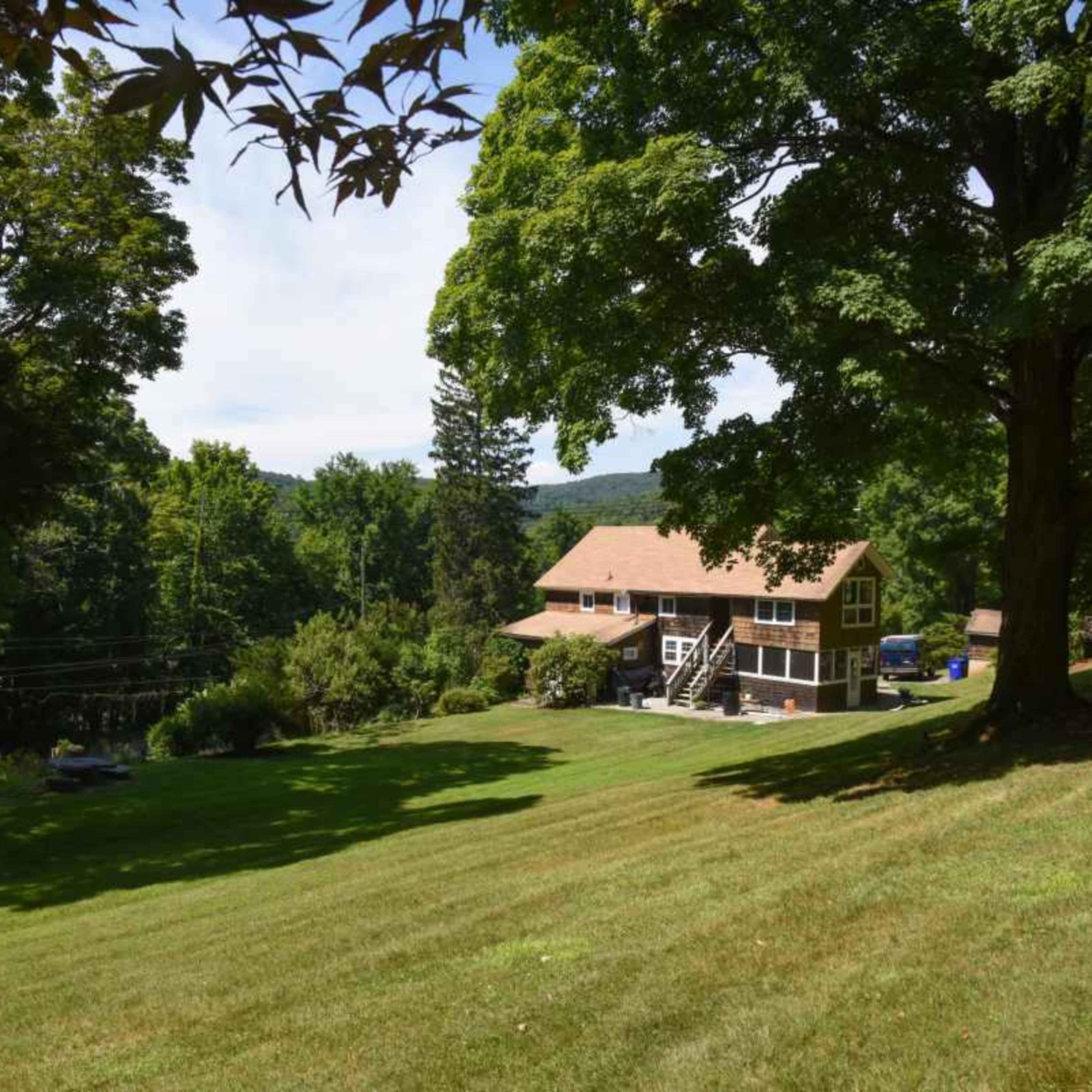 A large house with a brown roof is situated on a grassy slope surrounded by trees and hills.