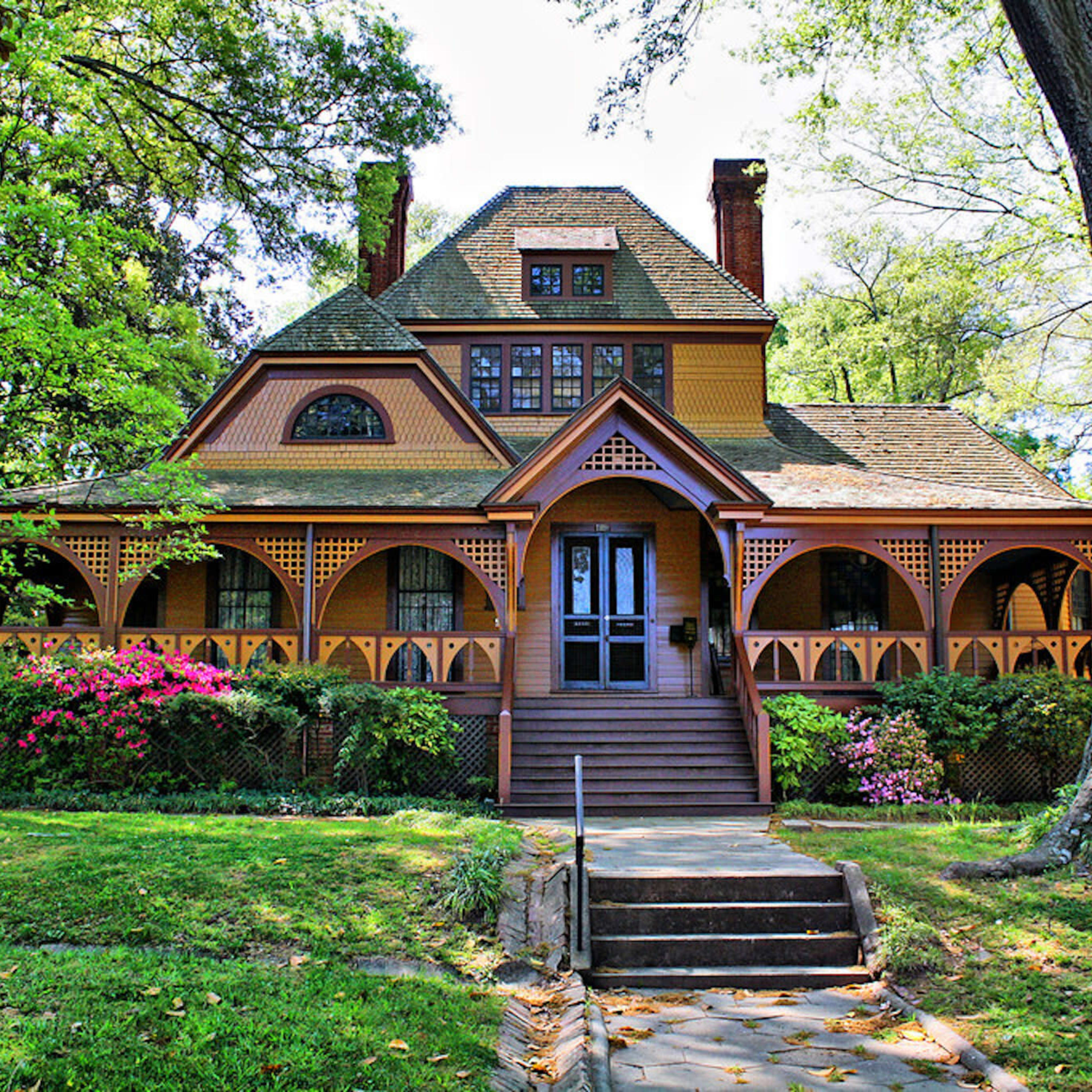 A large Victorian-style house with a wraparound porch and colorful flower beds sits surrounded by green trees and grass.