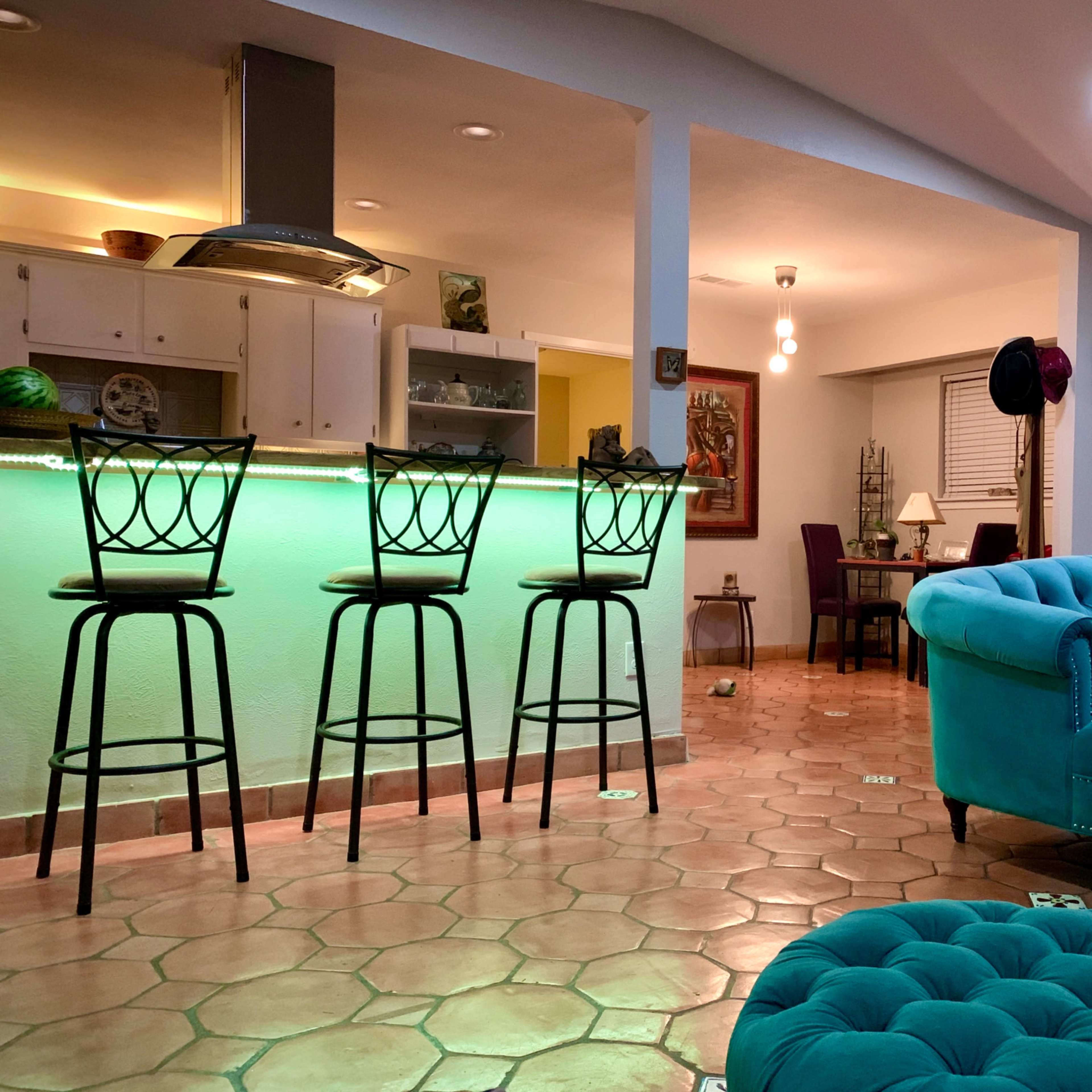 A brightly lit living space features a turquoise sofa, a modern bar with high stools, and a dining area in the background.