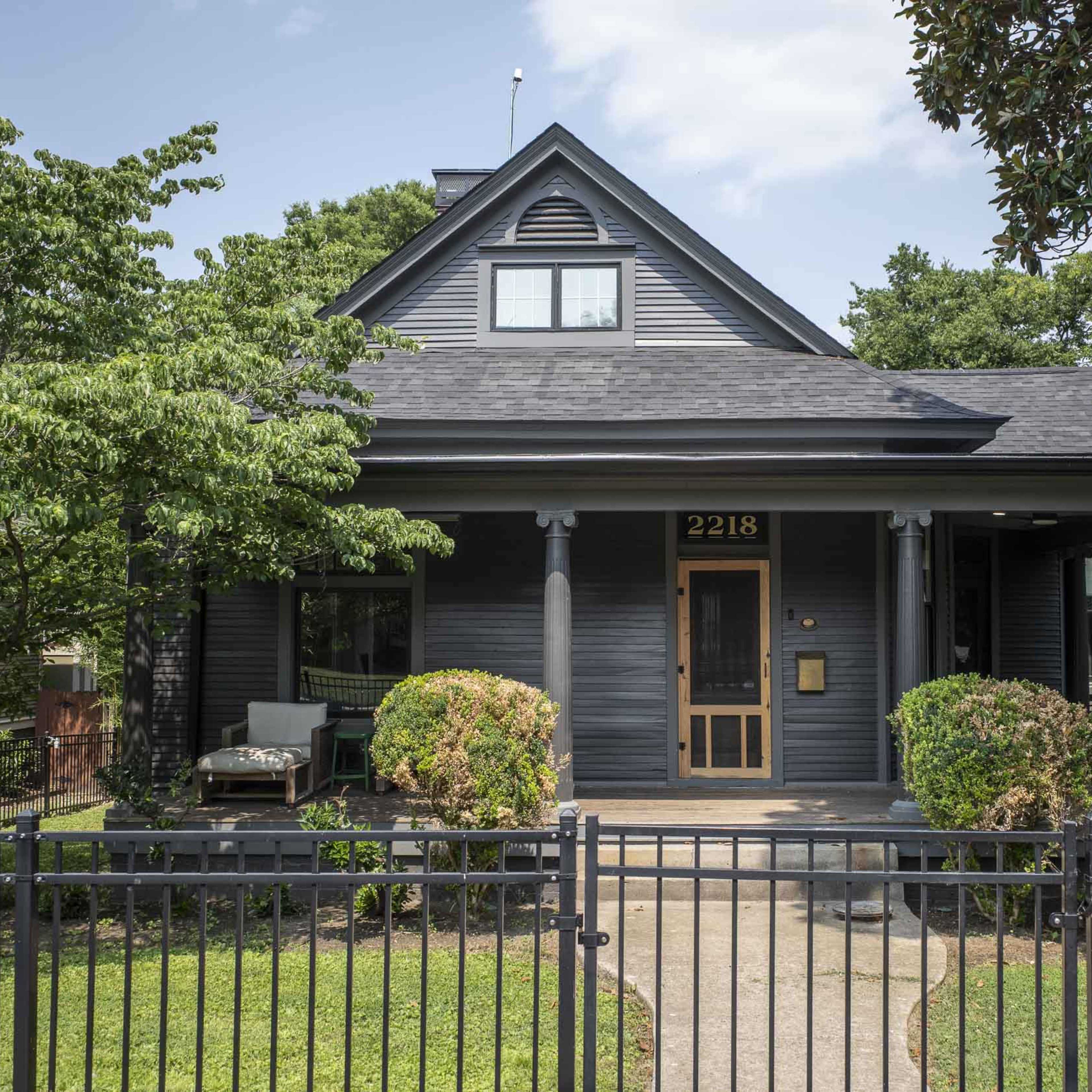 A dark-colored house with a front porch and landscaped greenery is set behind a metal fence.